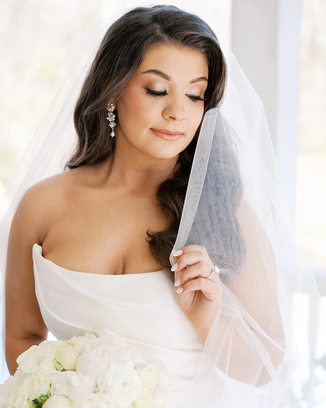 A bride with long dark hair, closed eyes, and elegant makeup, holding her veil delicately while wearing a white strapless wedding dress and silver earrings, standing near a window with natural light.