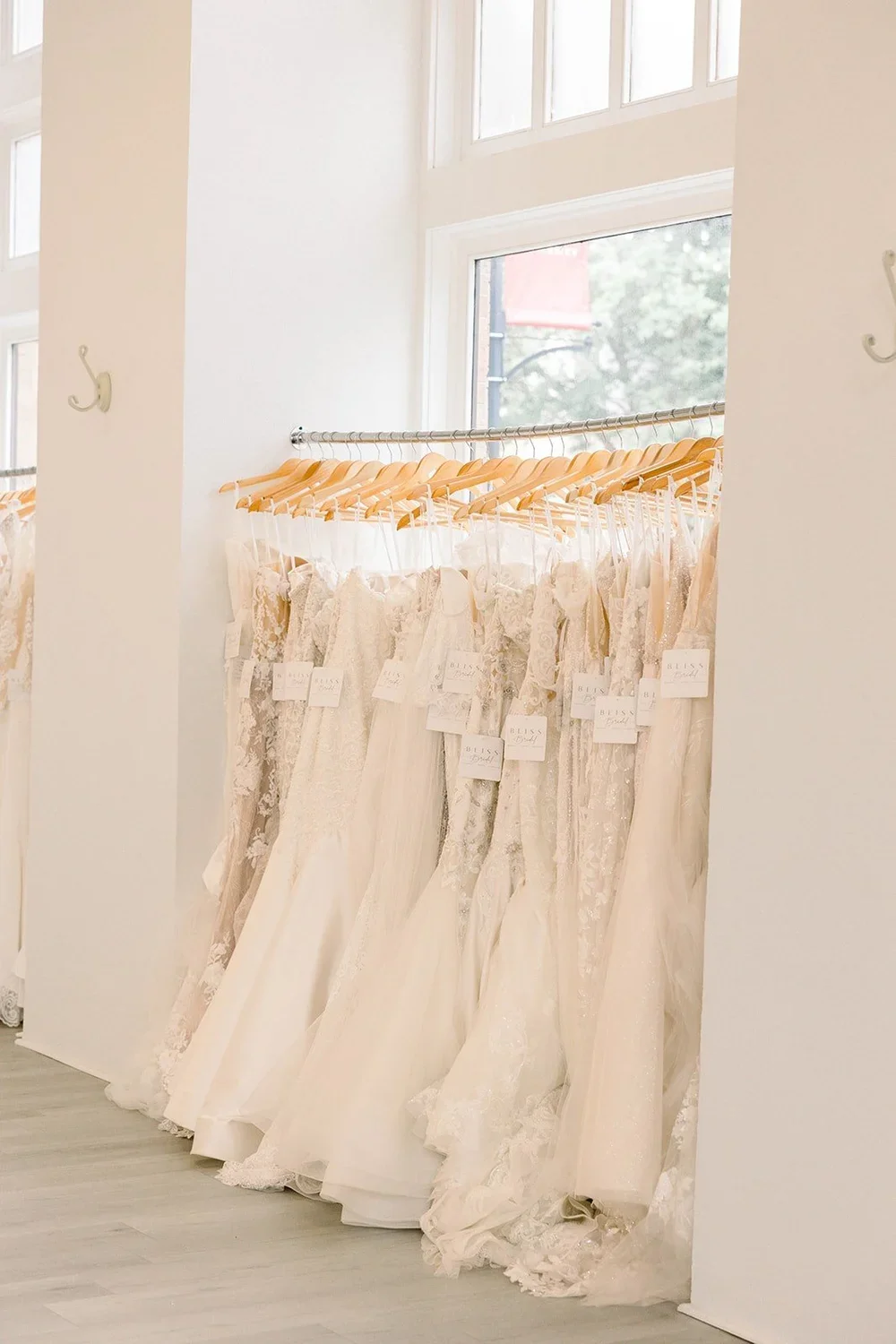 A row of white wedding dresses hanging on wooden hangers on a metal rack inside a bridal shop. The dresses feature lace and satin fabrics, and small tags are attached to each one. Natural light enters through large windows.