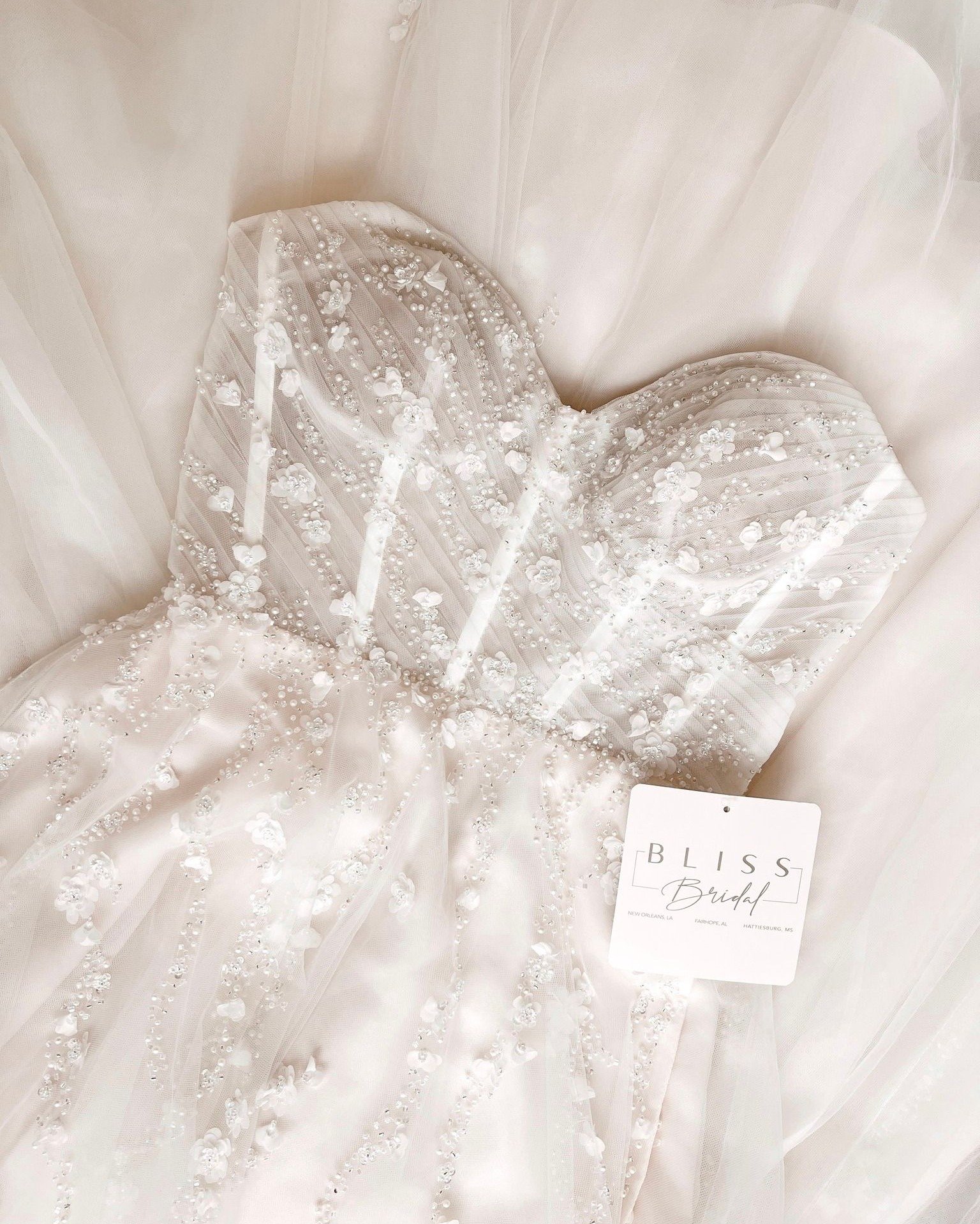 A white wedding dress with intricate bead and floral embroidery detailed with pearls and sequins, displayed on a light-colored surface with a tag reading 'BLISS Bridal' from New Orleans, LA, Faroupe, AL, Hattiesburg, MS.