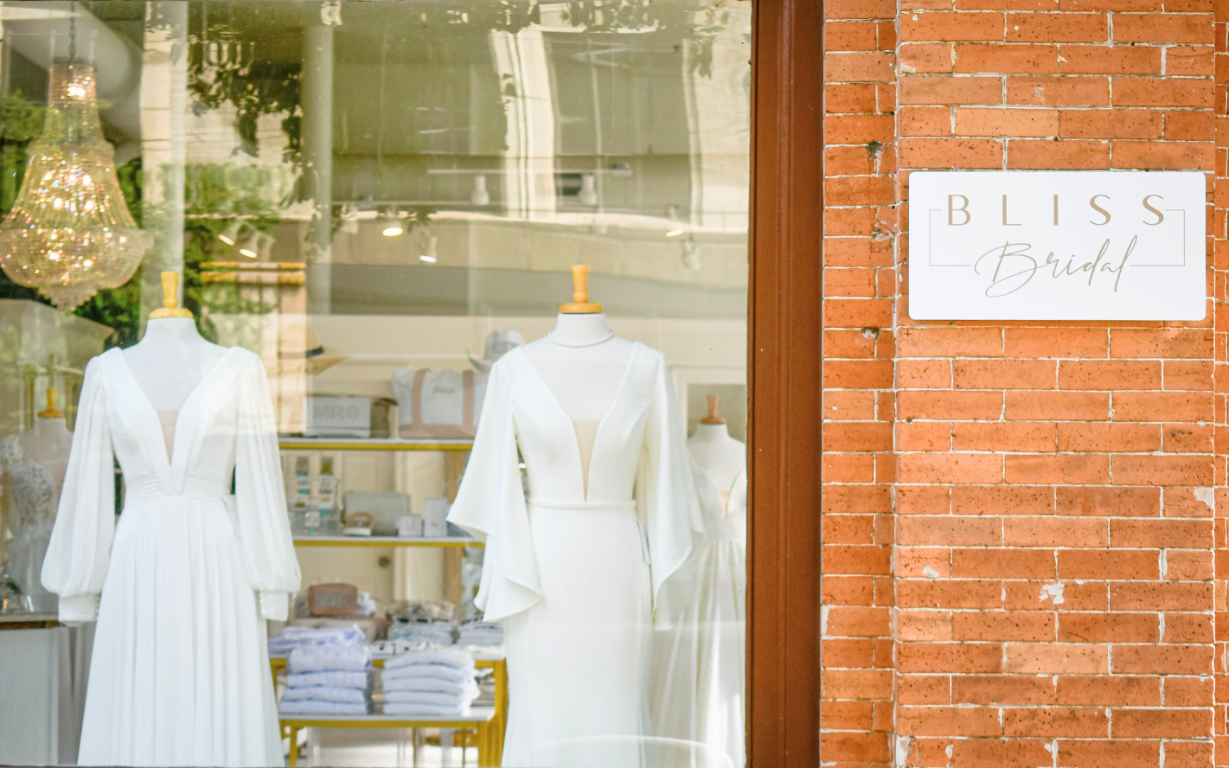 Storefront window display of white bridal dresses, with a sign that reads 'Bliss Bridal' on a brick wall outside.