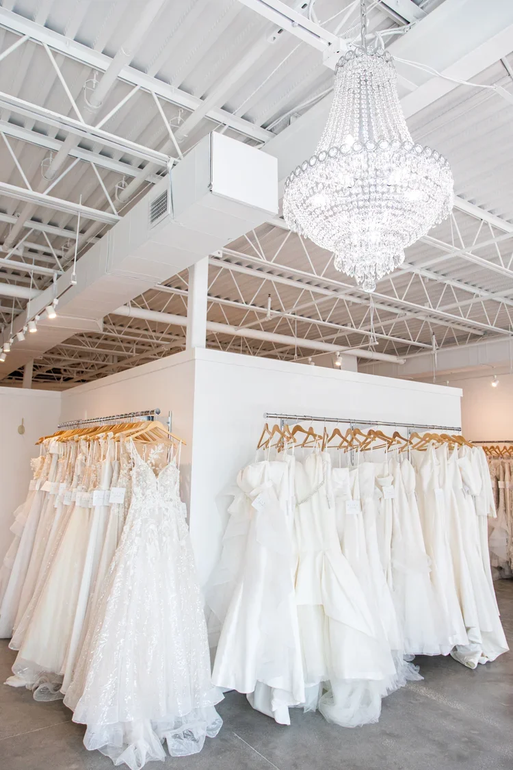 A store display with rows of white wedding dresses hanging on wooden hangers in a bridal boutique with a large crystal chandelier hanging from the ceiling.