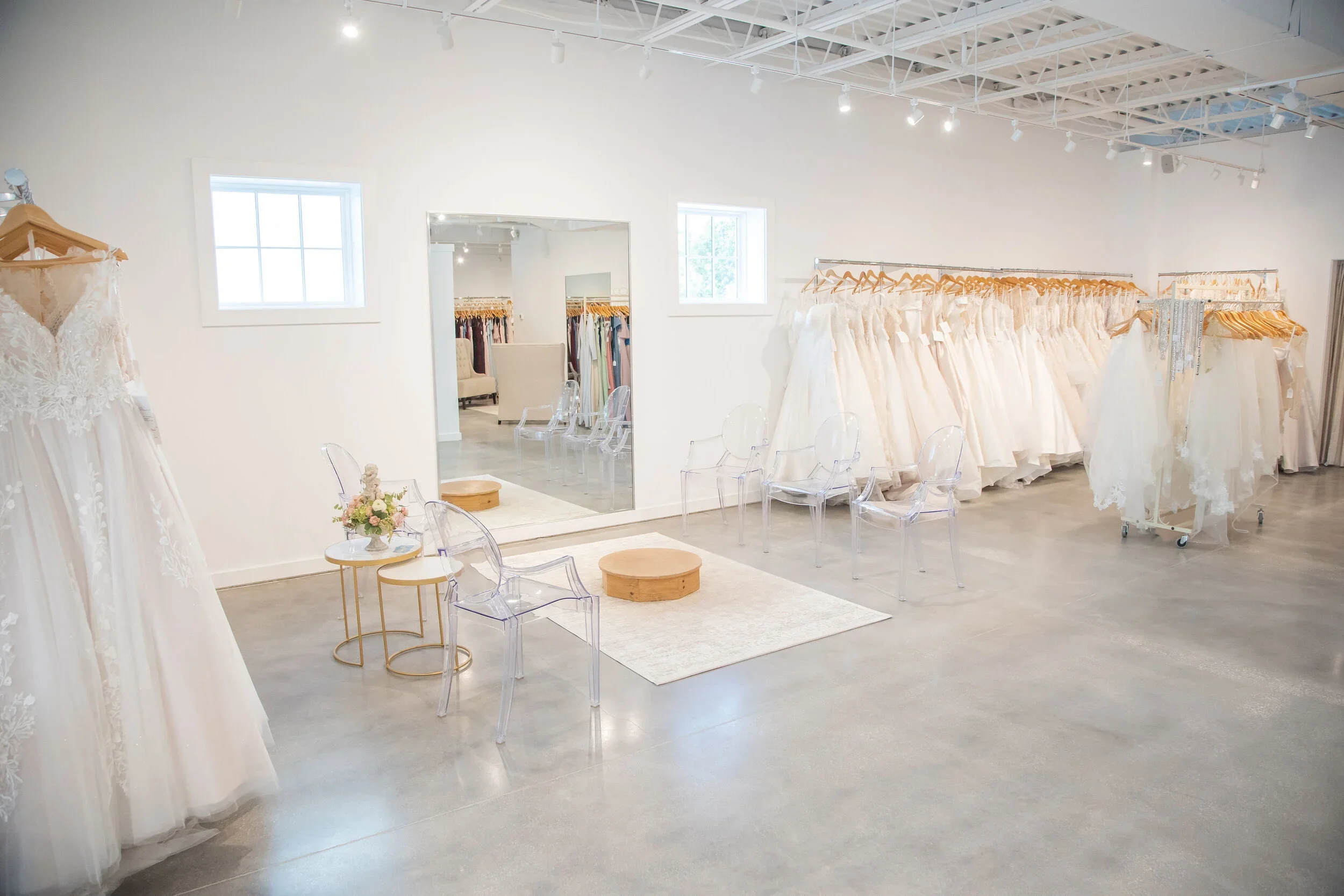 Wedding dress boutique with white walls, windows, and a variety of wedding gowns on racks, clear chairs, a mirror, and a small seating area.