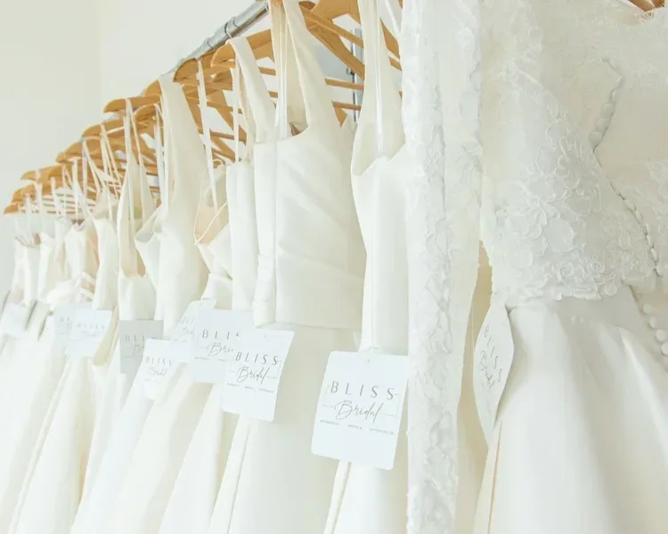 Several white bridal dresses hanging on a wooden rack, each with a tag attached.
