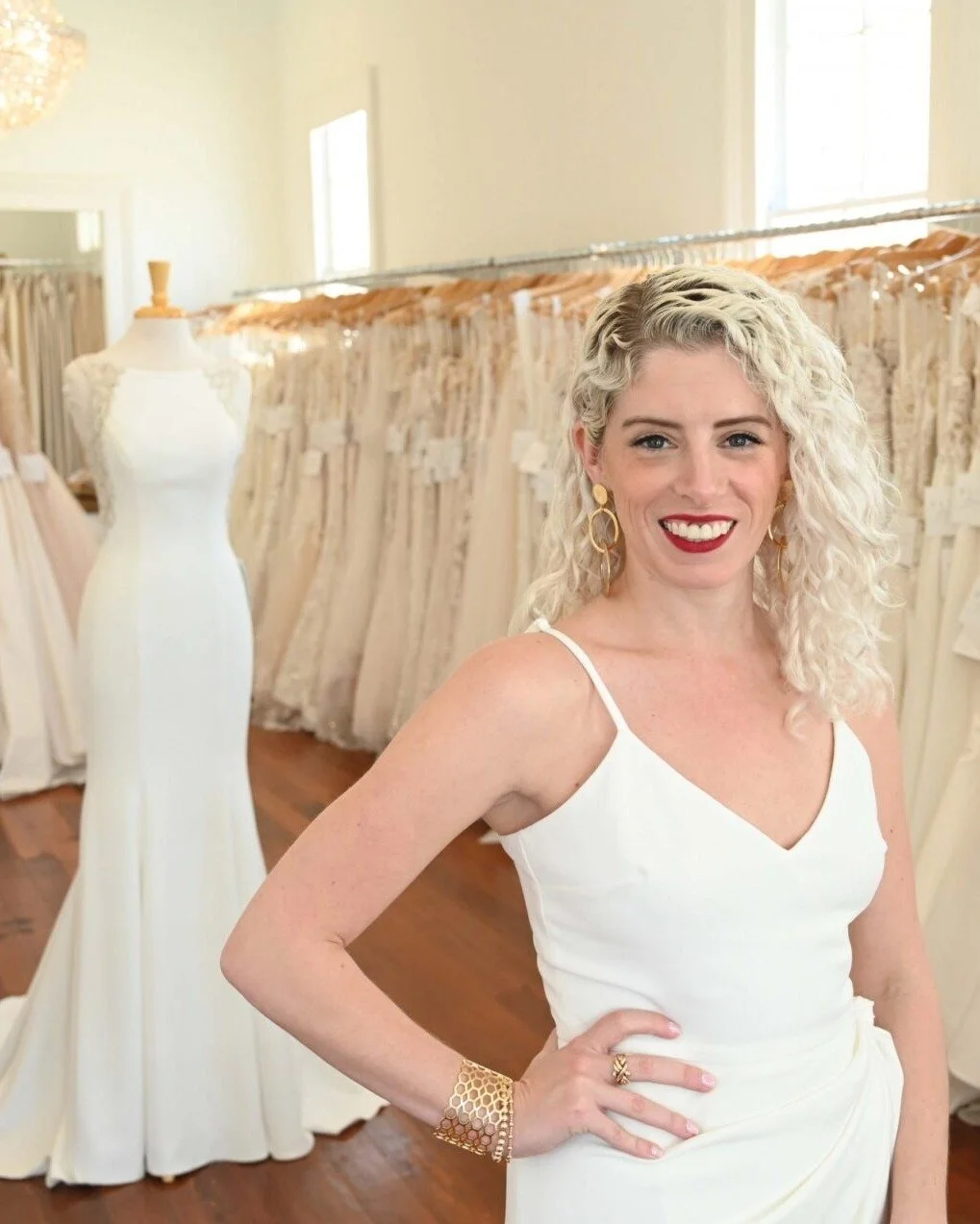 A woman with blonde curly hair and red lipstick wearing a white dress, standing in a bridal boutique with wedding dresses on racks behind her.