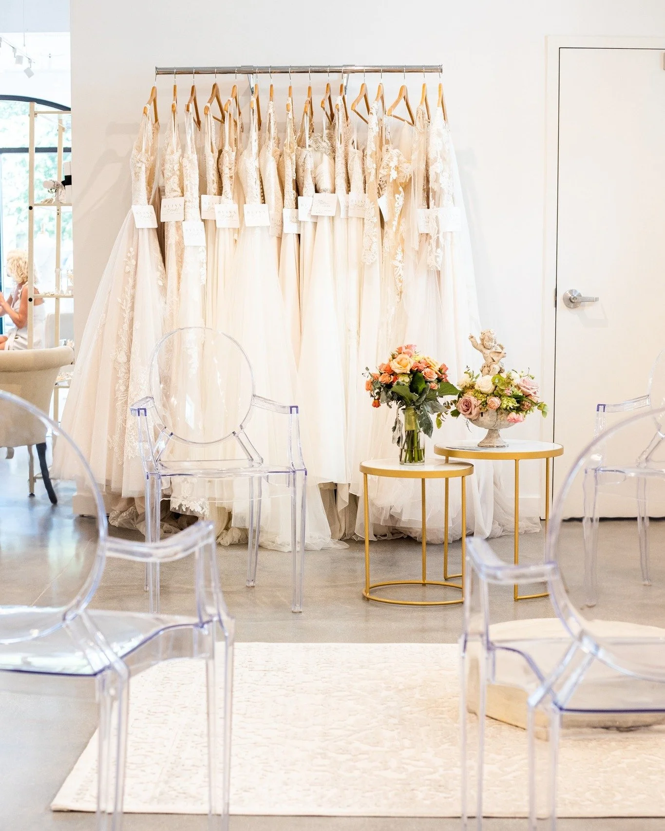Wedding dresses hanging on a rack in a boutique with clear ghost chairs and small gold tables with floral arrangements