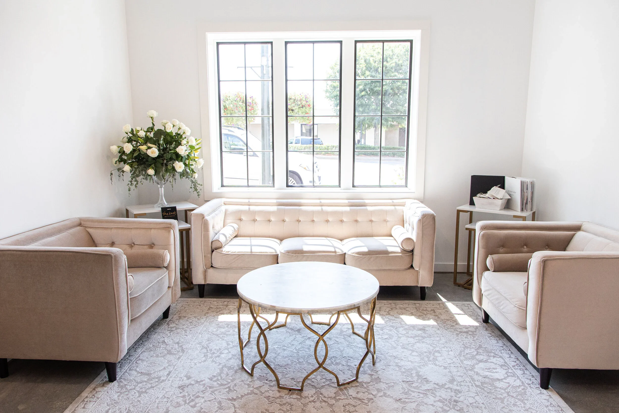 Bright living room with white walls, large front window, beige sofas, a white marble-top coffee table with gold legs, and side tables with a bouquet of white roses and papers.