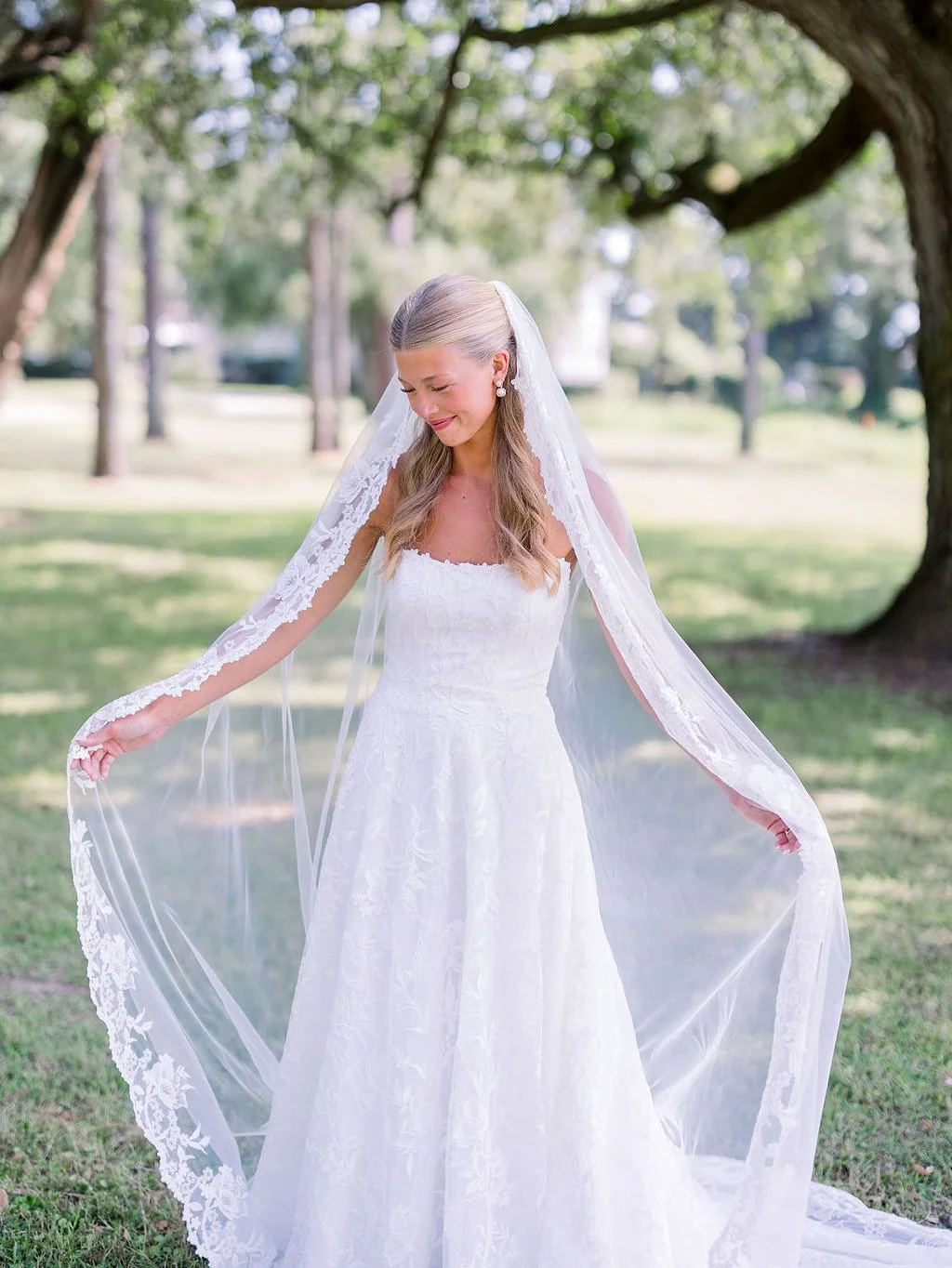 A bride in a white wedding gown with lace details, standing outdoors under a large tree with green leaves, smiling and holding the edges of her veil.