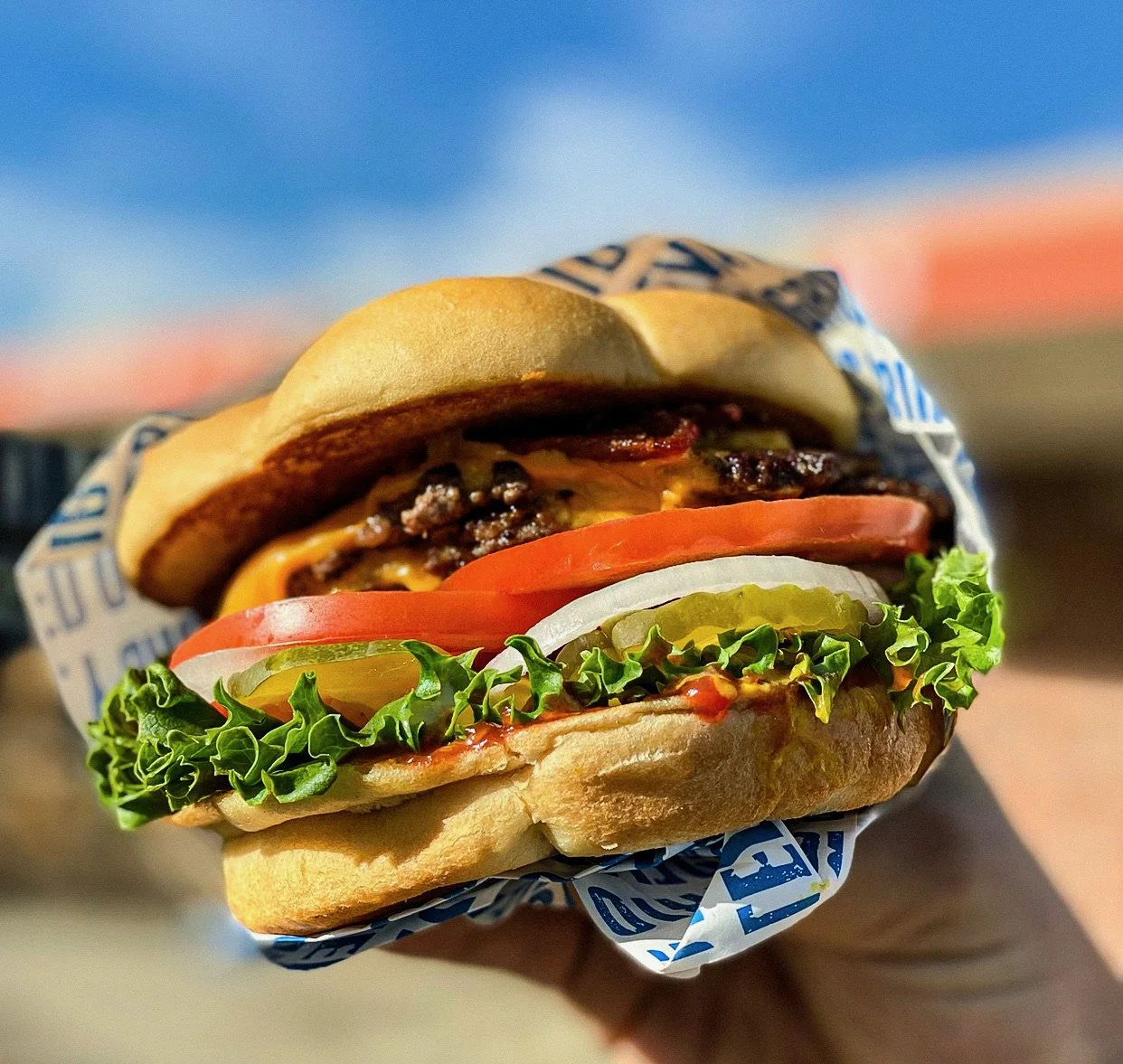 Close-up of a cheeseburger with lettuce, tomato, onion, pickles, cheese, and a beef patty, wrapped in blue and white paper.