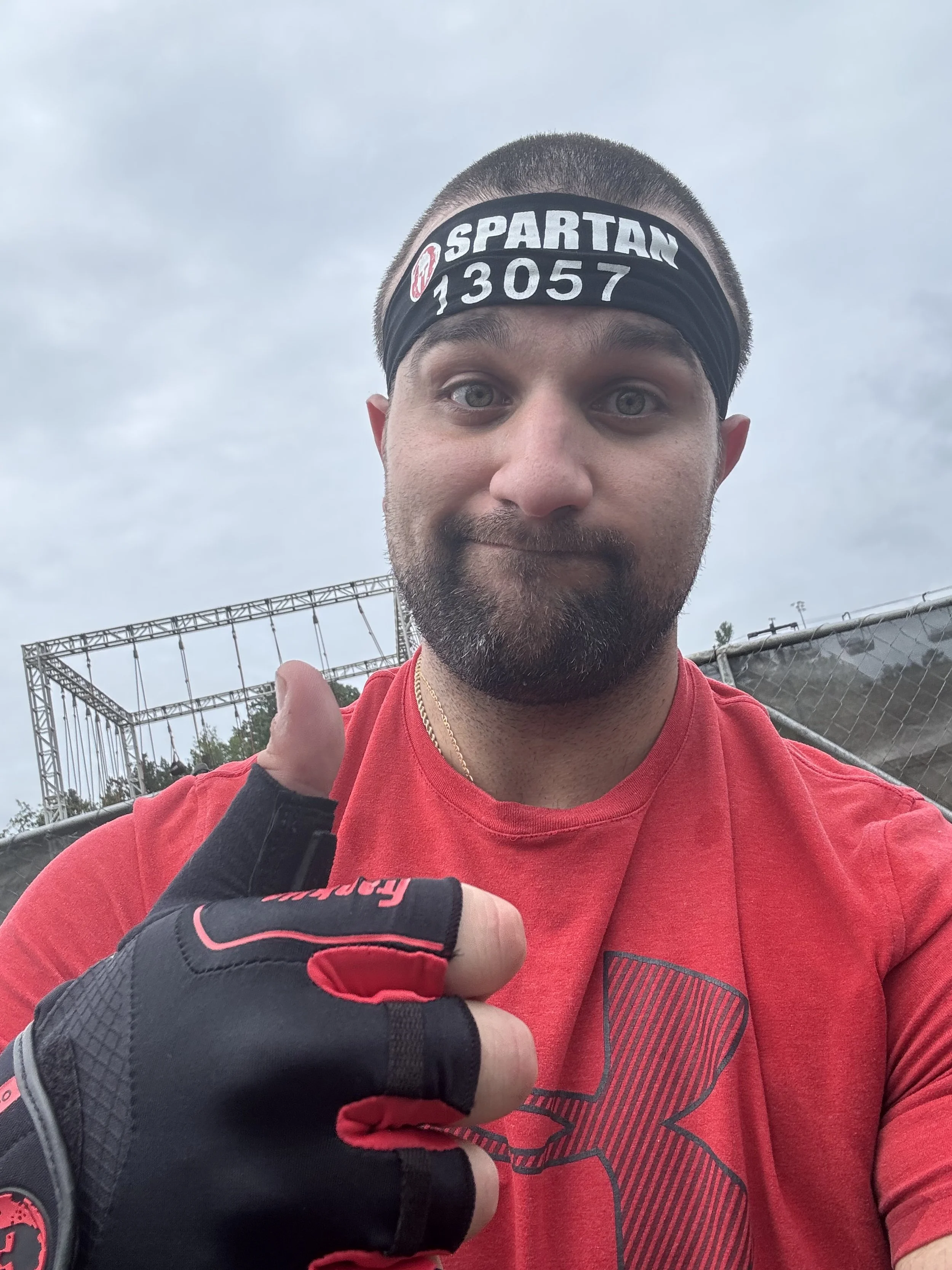 A man taking a selfie outdoors, wearing a black headband with white text that says "SPARTAN" and a number, red athletic shirt with a logo, and black and red fingerless gloves. He is giving a thumbs-up and has cloudy skies in the background.