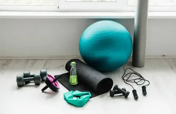 Fitness equipment including dumbbells, a resistance band, a yoga mat, a stability ball, a foam roller, a jump rope, and a water bottle on the floor near a window.