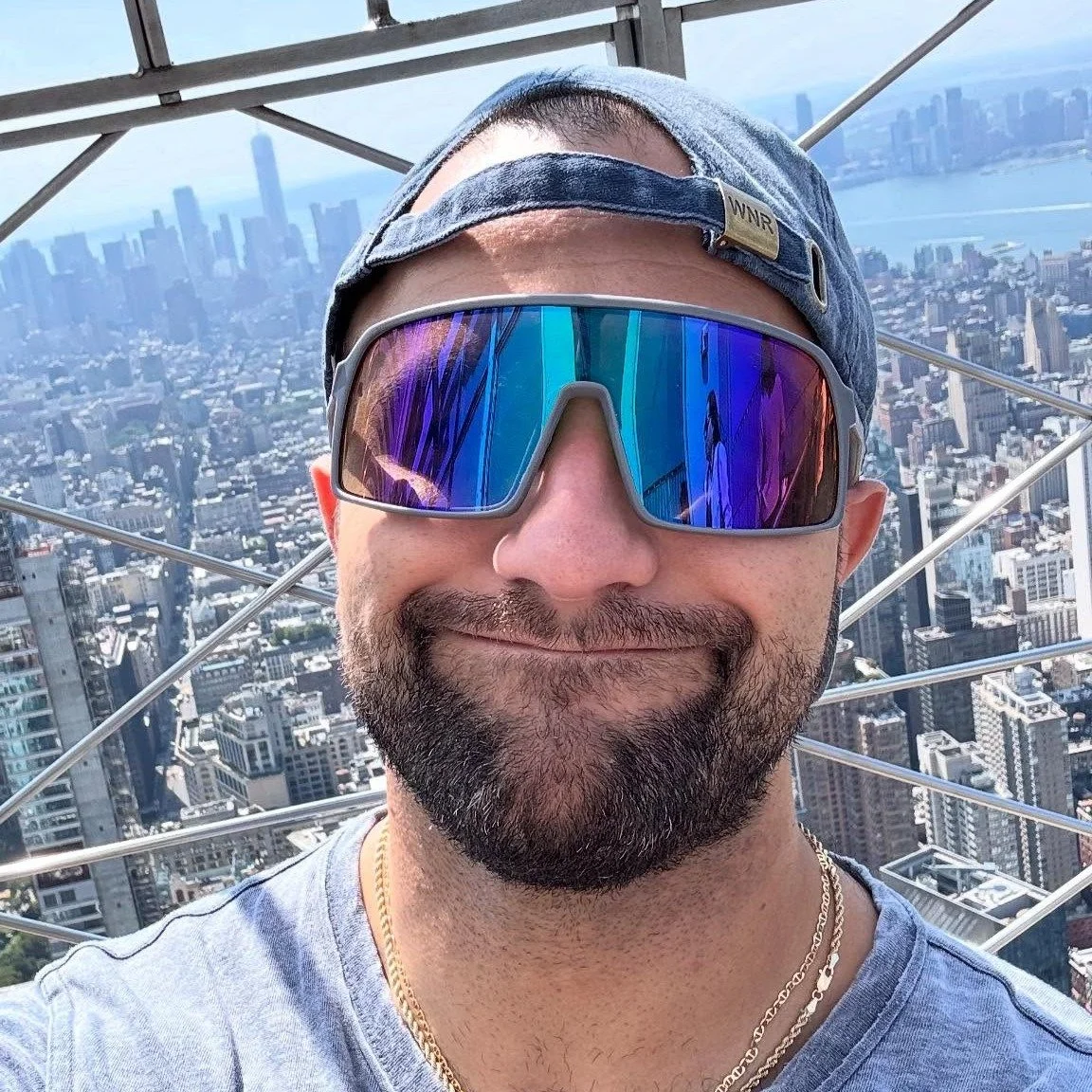 A man smiling and taking a selfie from a high observation point in New York City, with the city skyline and tall buildings in the background.