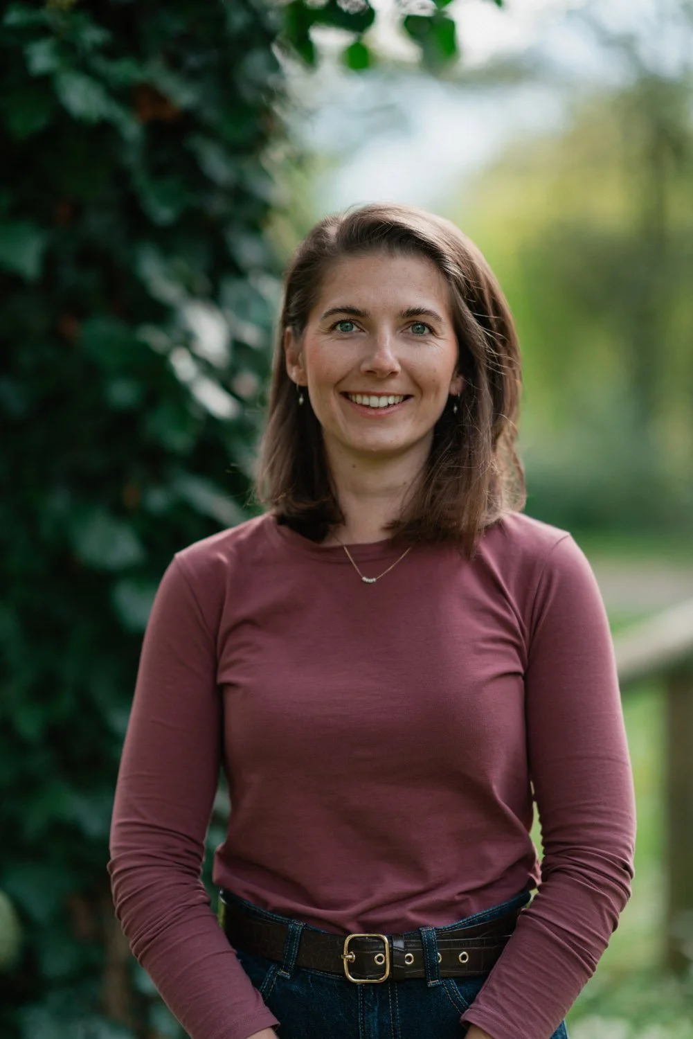 Junge Frau im Freien mit braunem Langarmshirt, lächelnd, hüfthoch, dunkelbrauner Haarkopf, grüner Hintergrund, wahrscheinlich im Park oder Garten.