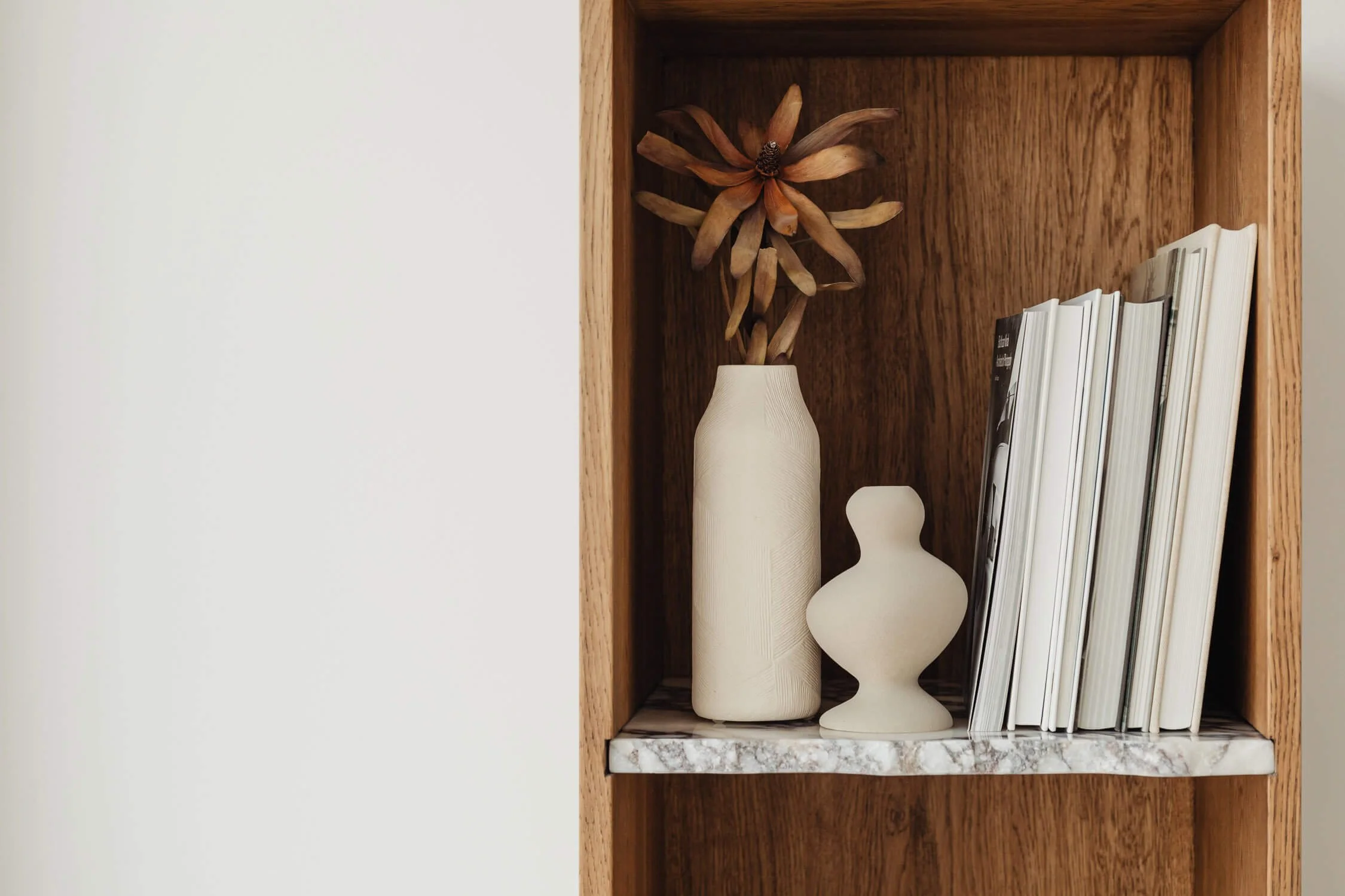 Holzregal mit weißen Vasen, einigen Büchern und einer getrockneten Blume aus Holz.