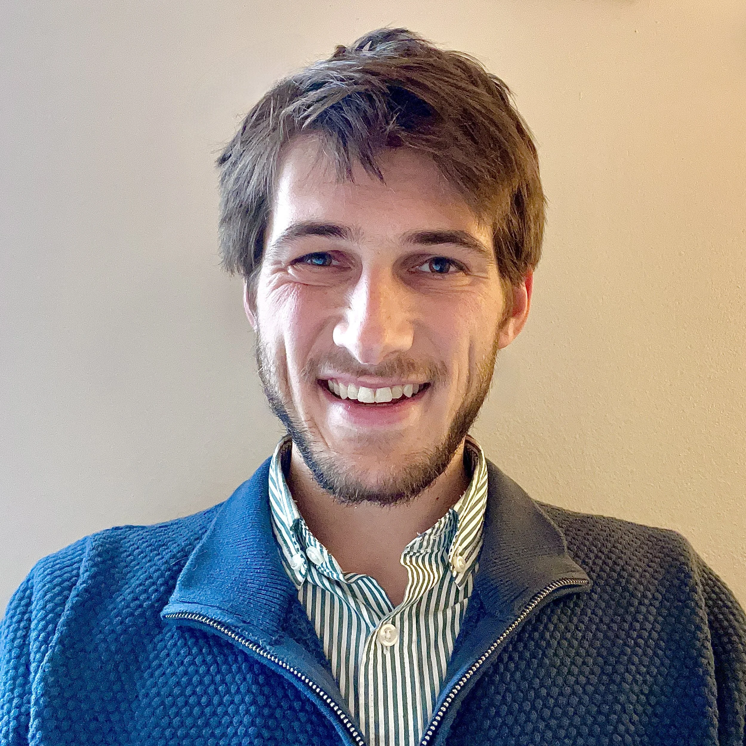 A young man with brown hair, blue eyes, and a beard, smiling, wearing a blue textured jacket over a green and white striped collared shirt, standing against a beige wall.