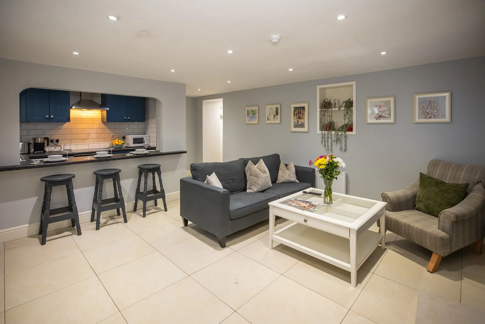 Living room with gray sofa, plaid armchair, white coffee table, and wall art. Open kitchen with blue cabinets, black countertops, and bar stools.