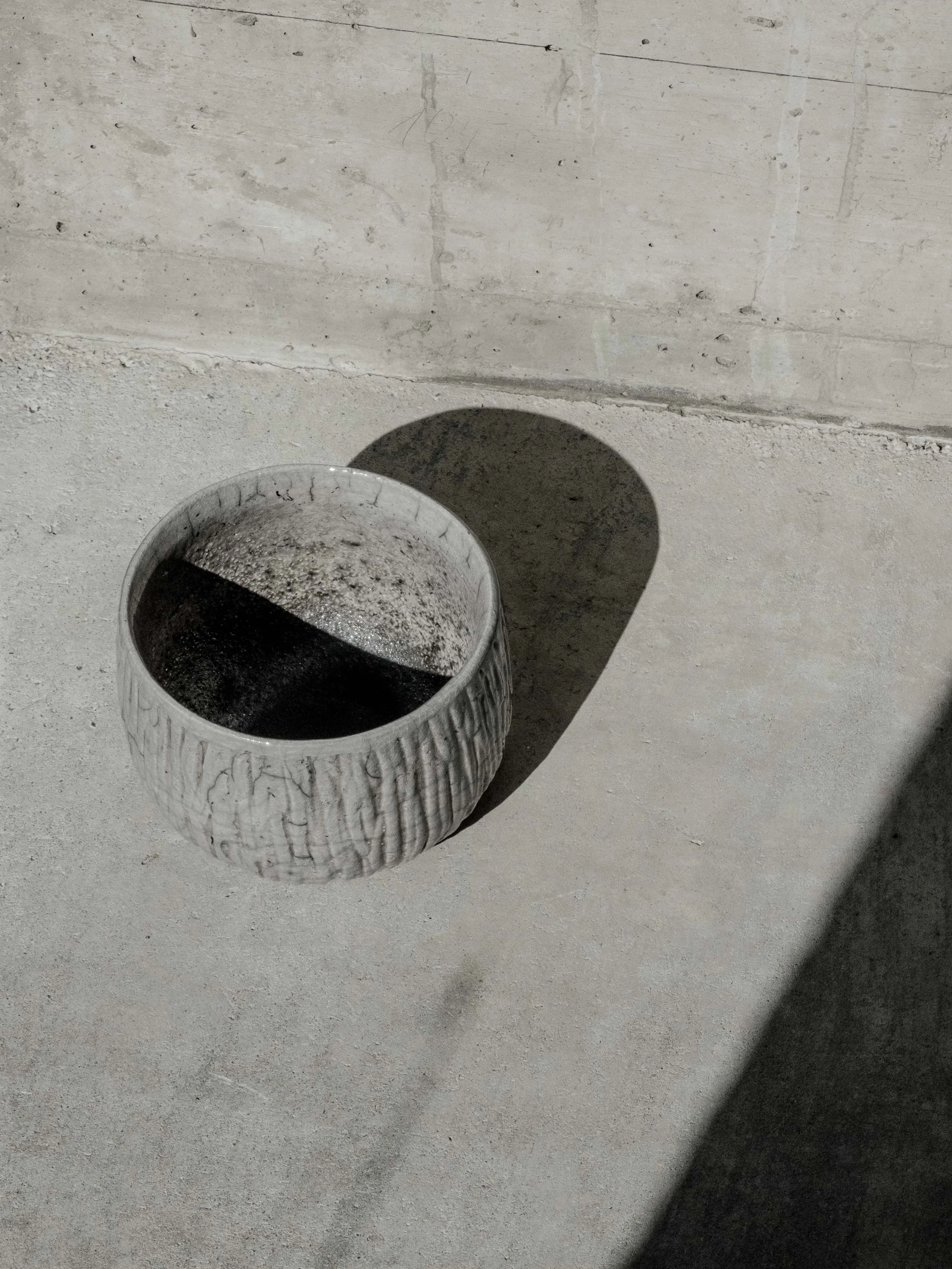 Ein Blumenkübel aus weißem Material auf einer Betonbodenfläche im Schatten. Der Schatten des Kübels ist sichtbar auf dem Boden.