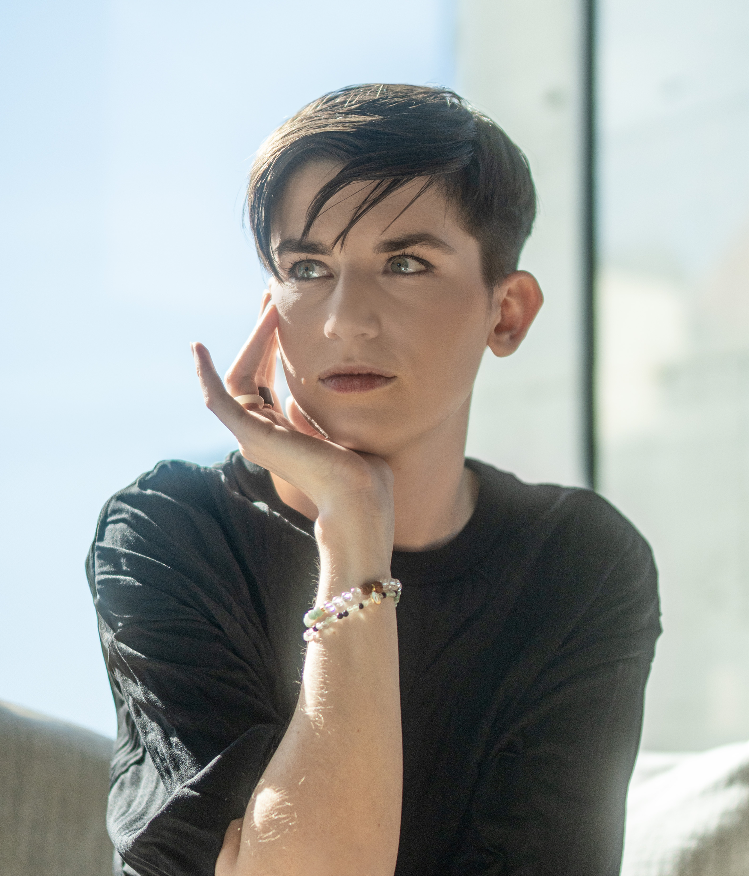 Junge Frau mit kurzen dunklen Haaren, trägt Armbänder und hat eine Hand am Kinn, schaut nach oben, im Hintergrund ein Fenster bei Tageslicht.