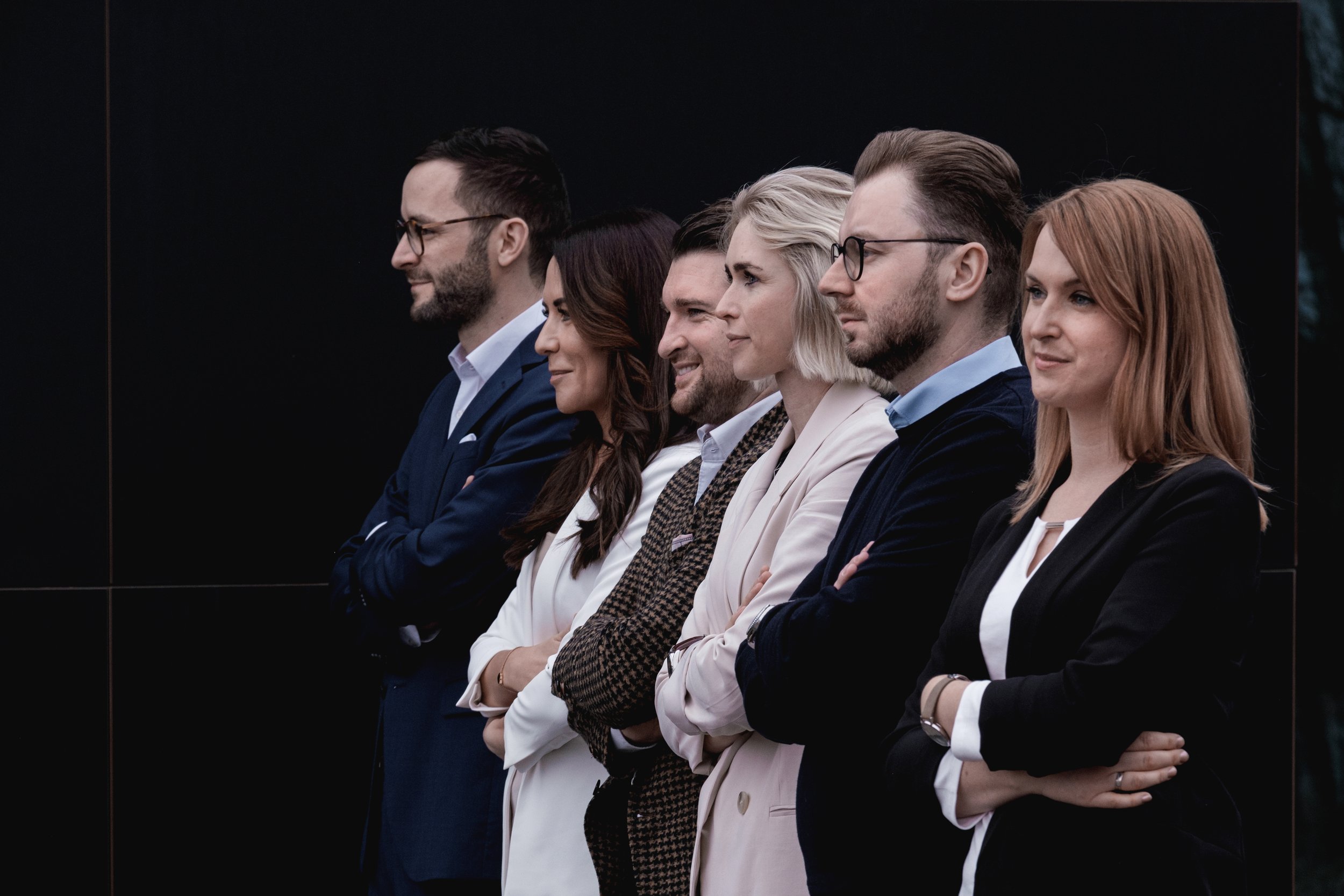 Sechs professionelle Menschen stehen in einer Reihe, mit verschränkten Armen, vor einem schwarzen Hintergrund, in einem formellen Umfeld.
