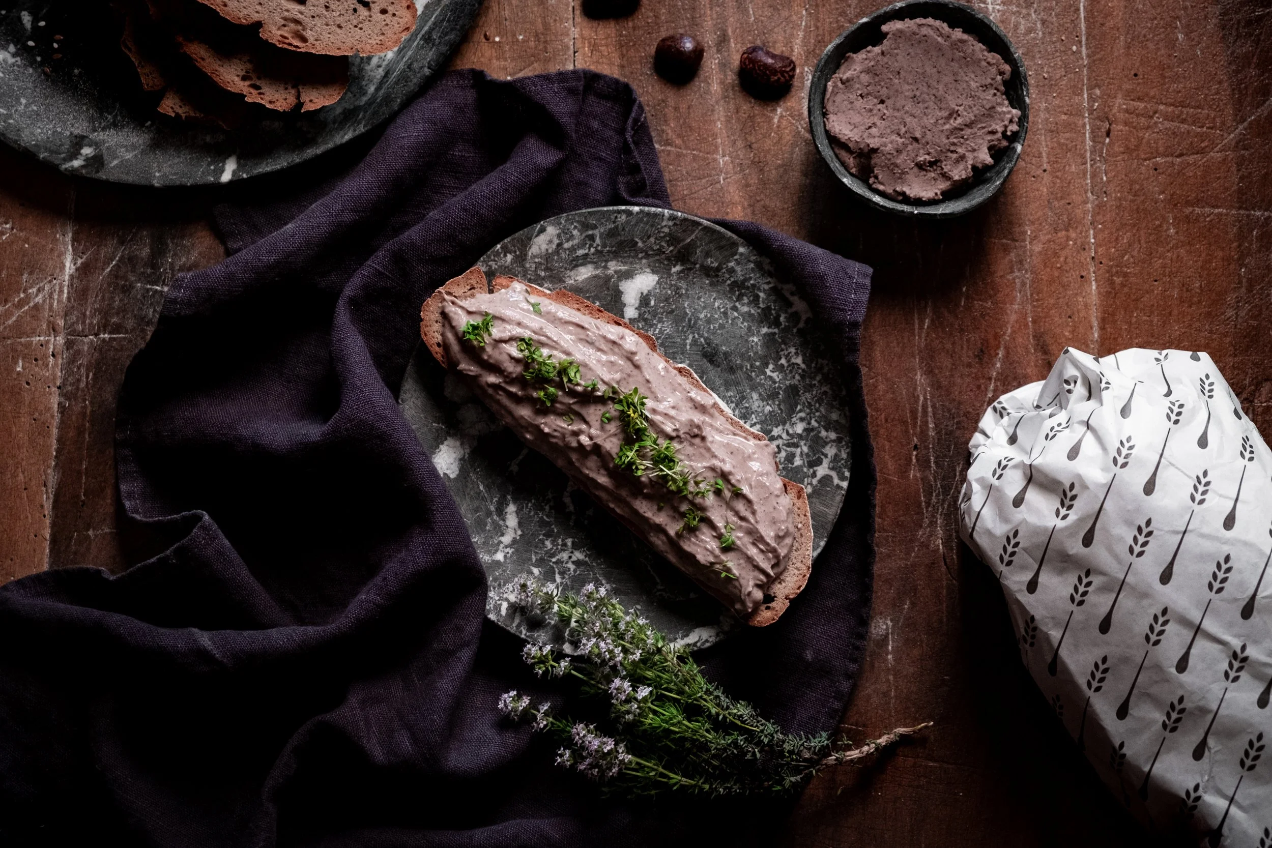 Brot mit Aufstrich und frische Kräuter auf einem dunklen Tuch, daneben ein Tuch mit Muster und helle Verpackung, auf einem Holztablett, im Hintergrund Tannenzweige mit kleinen lila Blüten.