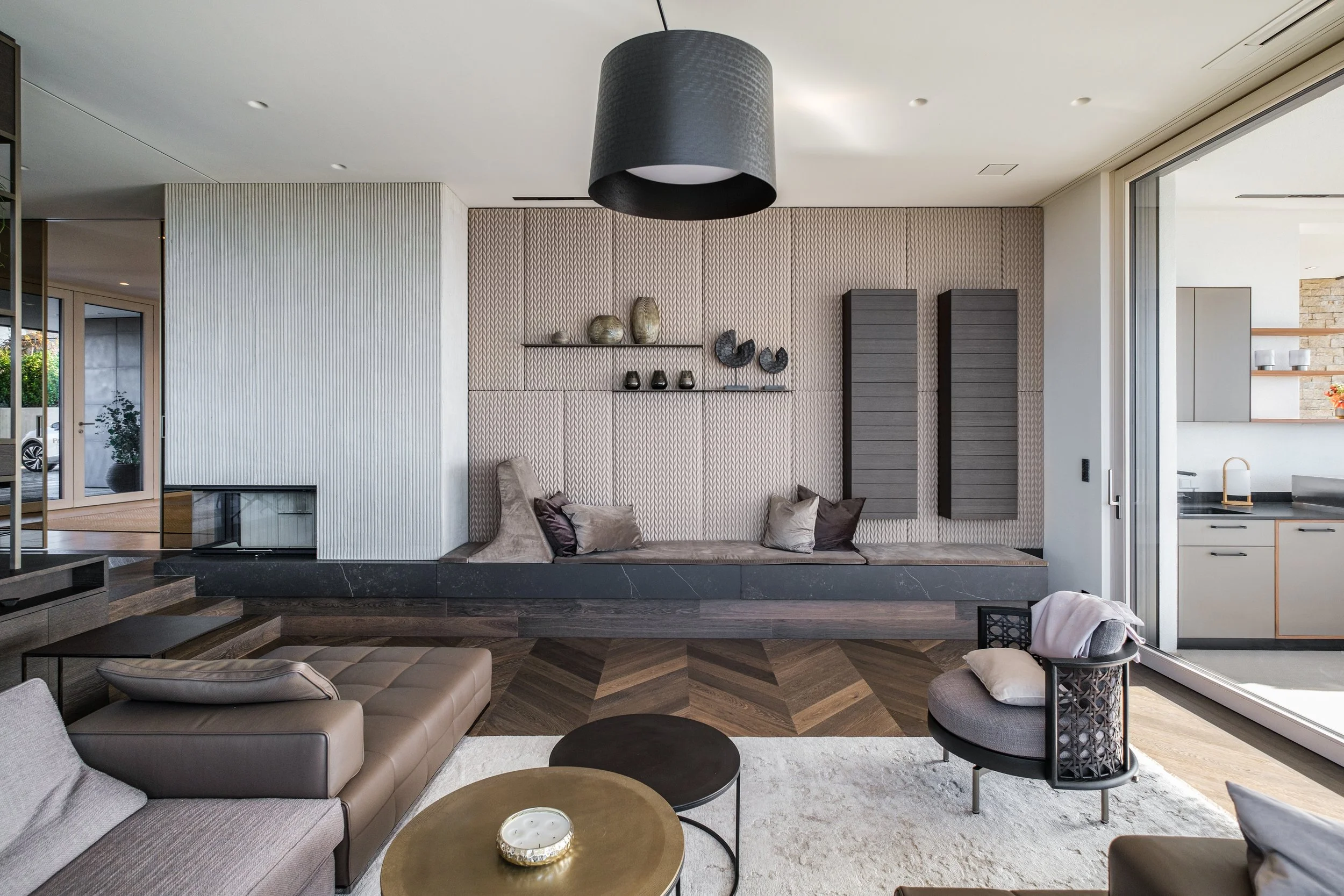 Modernes Wohnzimmer mit grauen und beigen Möbeln, dunklen Holzboden, Wanddeko und großem schwarzen Lampenschirm an der Decke, große Fenster und Zugang zur Küche im Hintergrund.