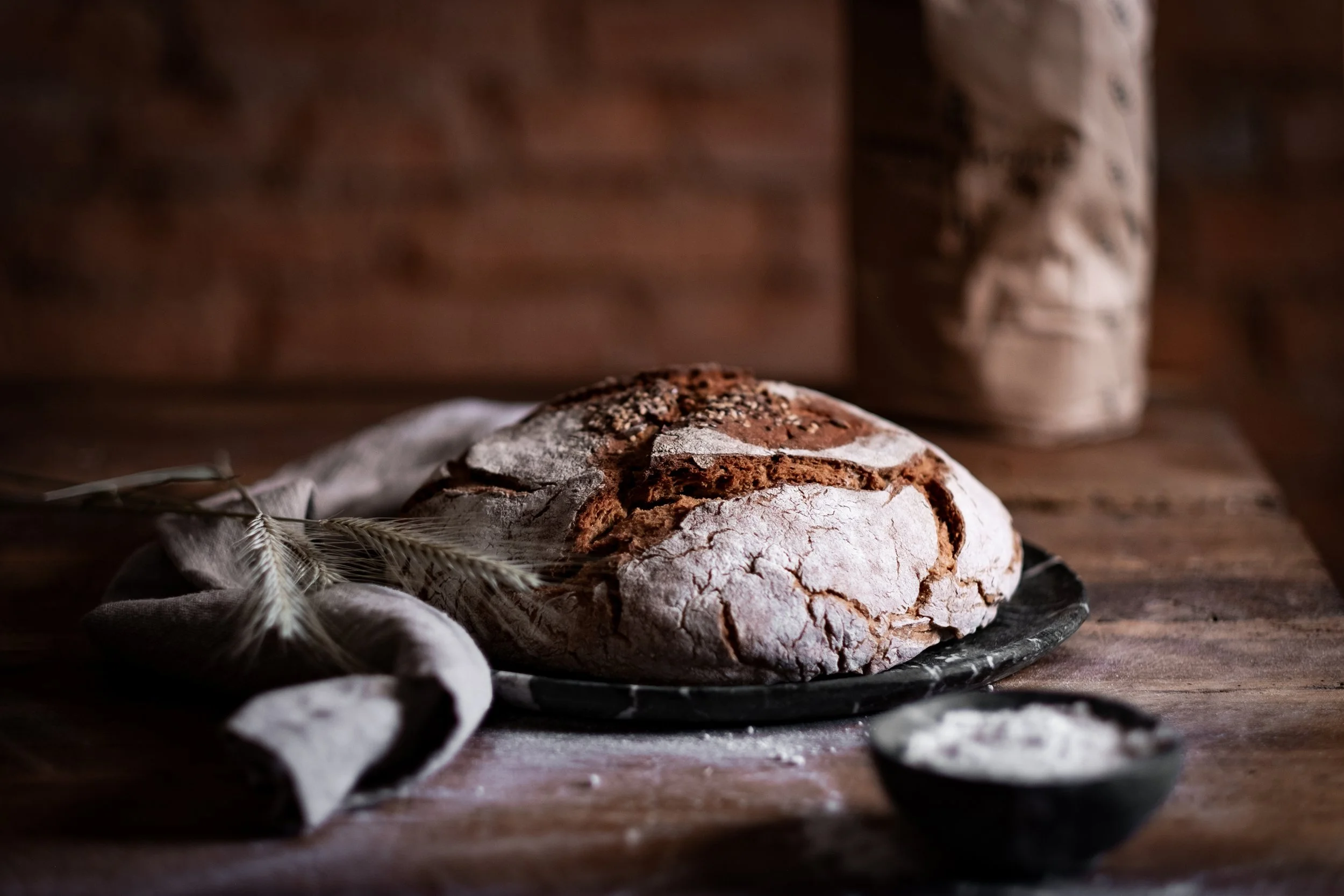 Ein rustikales Brot auf einem dunklen Teller, umgeben von Mehl und Weizenähren, auf einem holzfarbenen Tisch
