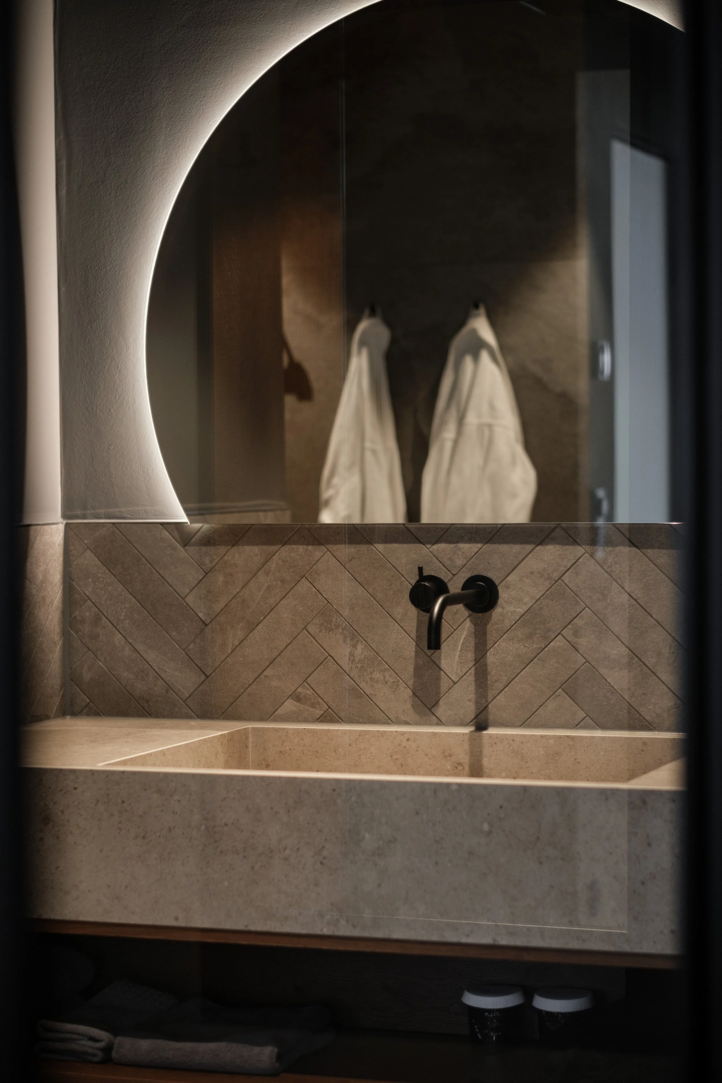 Ein Badezimmerwaschbecken mit dunklem Wasserhahn, einem Spiegel mit Beleuchtung, der eine Person mit weißen Hosen reflektiert, und dahinter eine dunkle Wand. Unten im Schrank befinden sich Handtücher, Toilettenpapier und zwei Papiertassen.