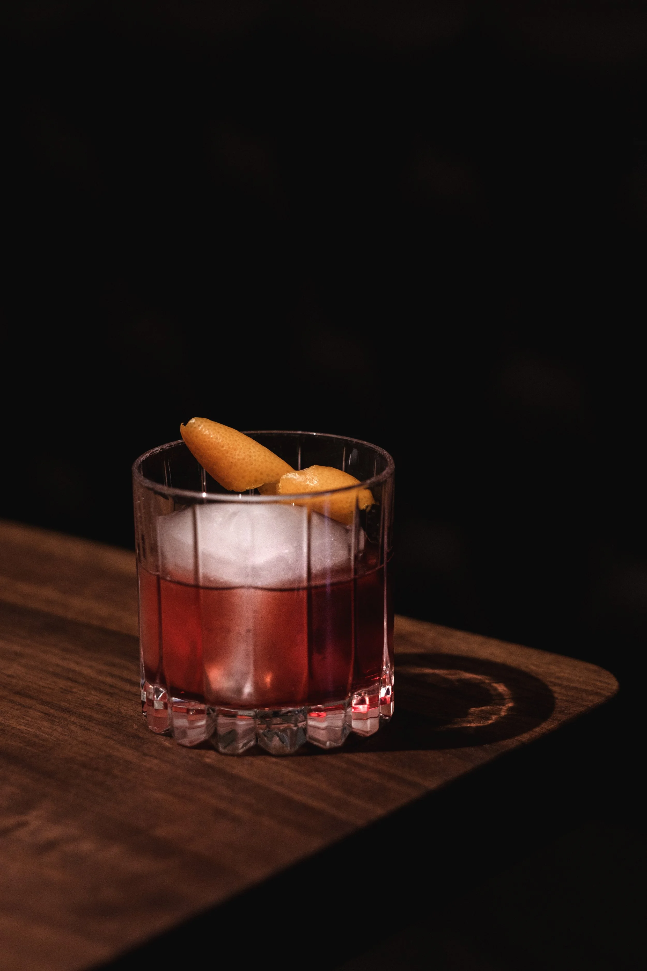 Ein Glas mit rot gefärbtem Getränk und einer Zitronenzeste als Garnitur, steht auf einem Holztisch vor dunklem Hintergrund.