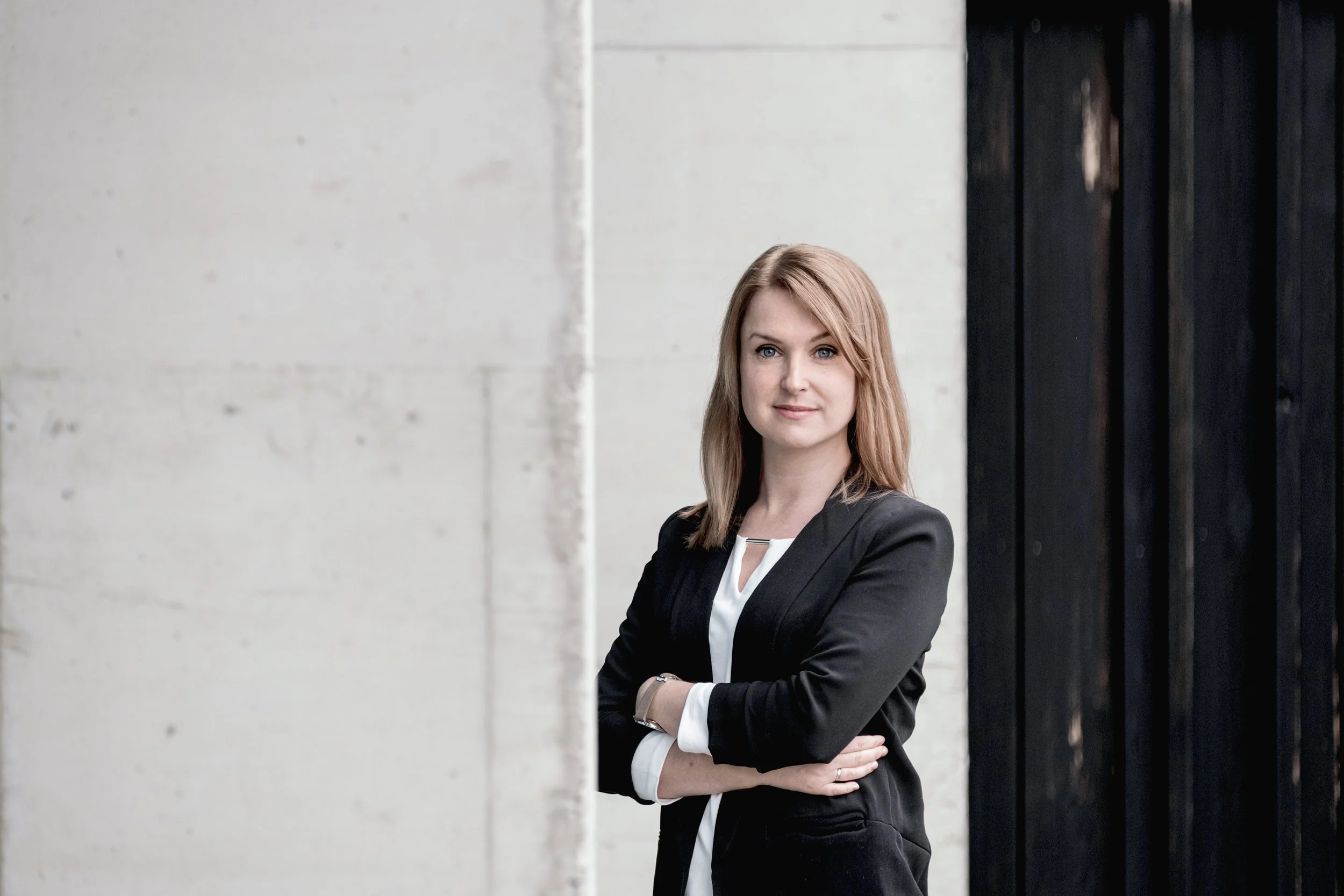Eine Frau mit roten Haaren, in einem schwarzen Blazer, die in die Kamera schaut. Sie steht vor einer weißen Wand und einer schwarzen Tür.