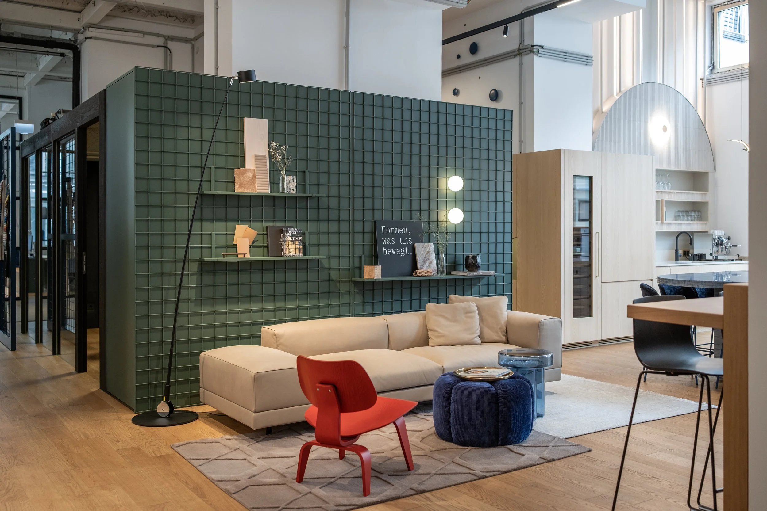 Interior des modernen Wohnraums mit grauer Couch, rotem Kinderstuhl, weißen Teppich, grüner Raumtrennwand mit Dekorationen und Büchern, Holzfußboden, Küche im Hintergrund, große Fenster, helle Beleuchtung.