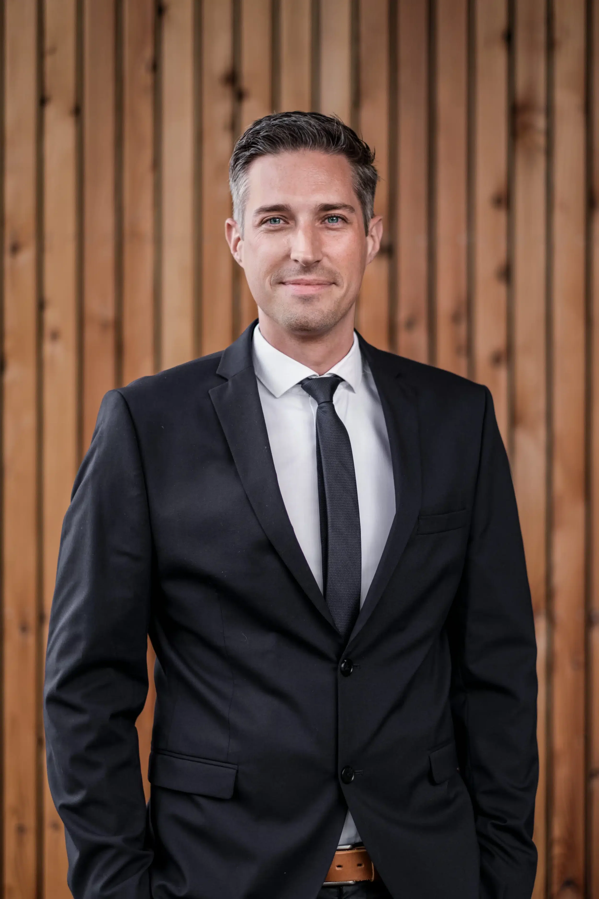 Ein Mann in einem schwarzen Anzug und Krawatte vor einem Holzwand. Er hat kurze dunkle Haare, blaue Augen und lächelt leicht.