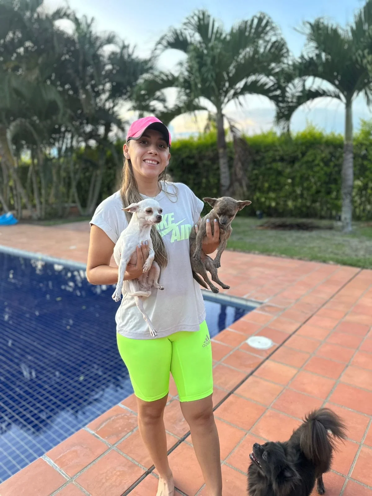 Una mujer con camiseta blanca, pantalones cortos verdes y gorra rosada sostiene dos chihuhuas perros pequeños, uno blanco y uno gris, junto a una piscina, mientras un perro más pequeño negro se encuentra en el suelo.