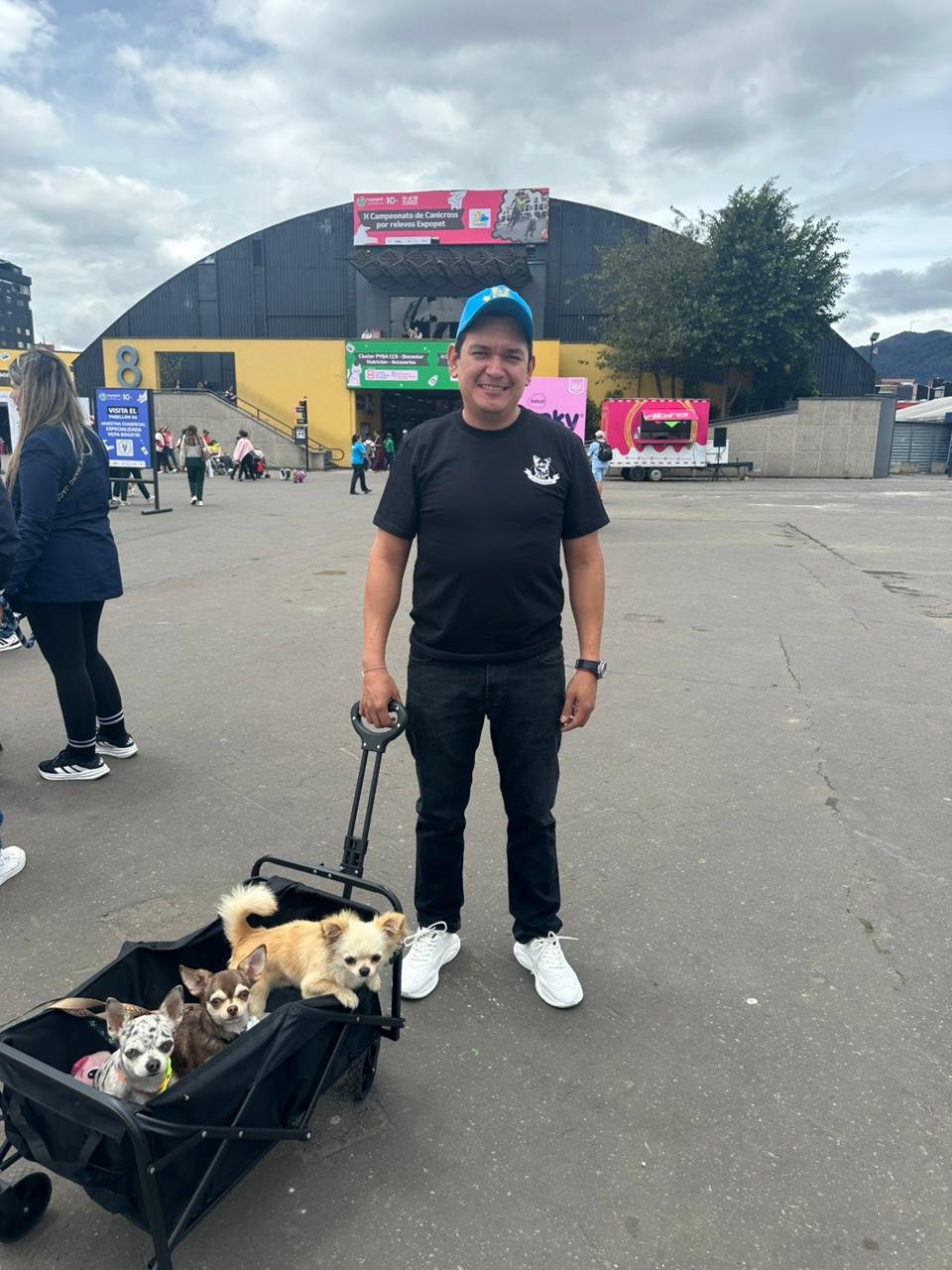 Hombre en camiseta negra y pantalones oscuros, con gorra azul, sonriente, de pie en un parque con cuatro perros pequeños en una carriola y otros perros en el fondo.