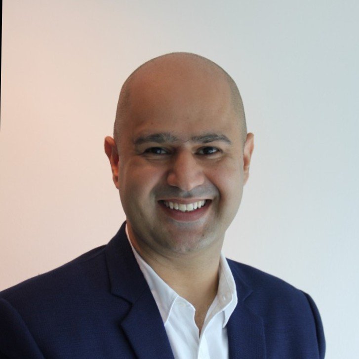 A smiling man with a bald head, wearing a dark blazer and white shirt, standing against a plain light-colored background.