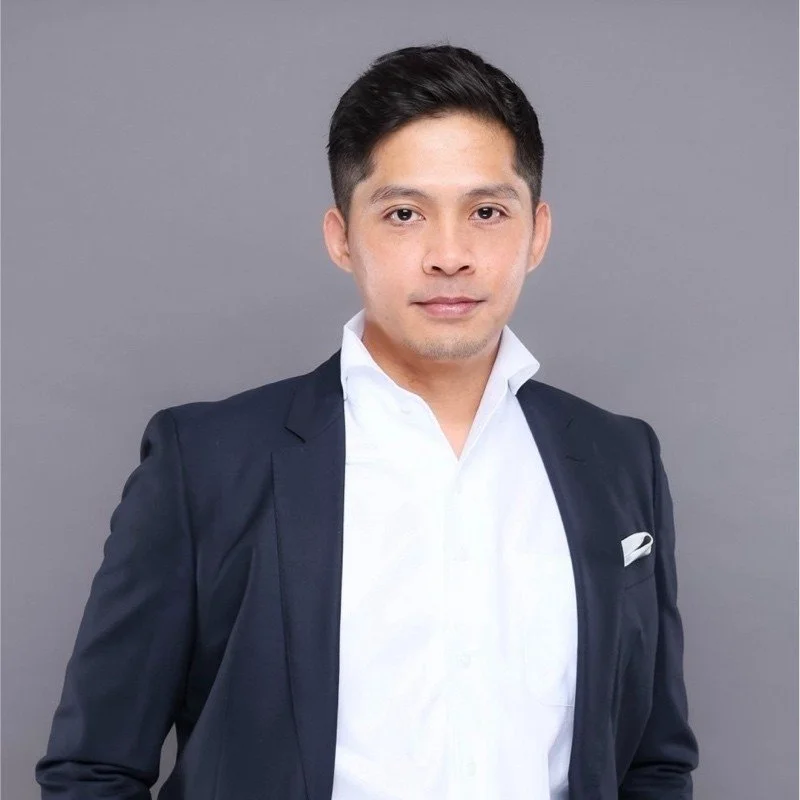 A man in a navy blazer and white shirt standing against a plain gray background.