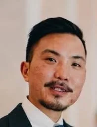 A man with short black hair and a mustache, wearing a dark suit and white shirt, smiling indoors.