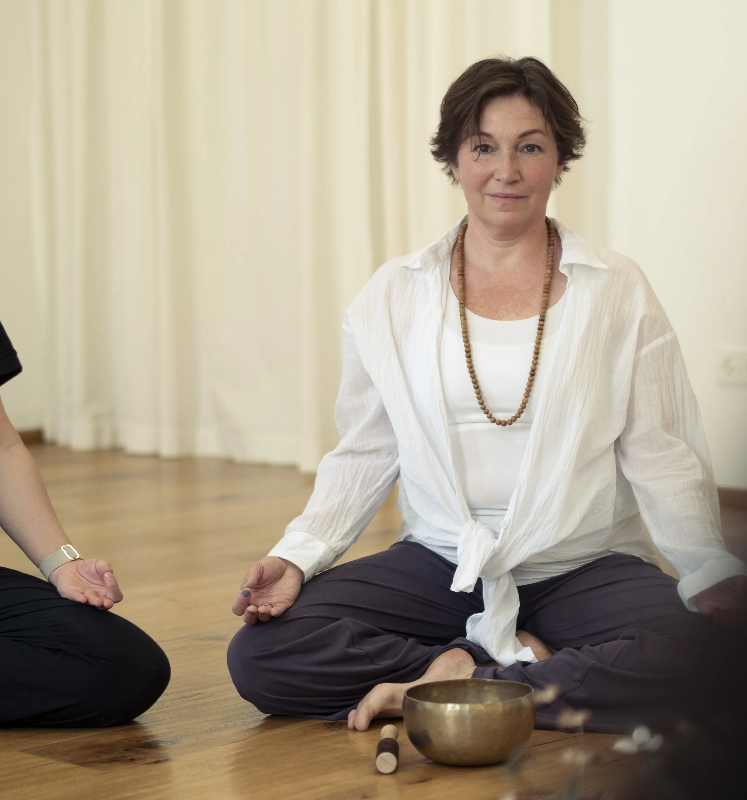 Zwei Menschen sitzen auf dem Boden und meditieren. Sie halten ihre Hände in einer meditativen Pose, und es sind Klangschalen und Meditationszubehör um sie herum.