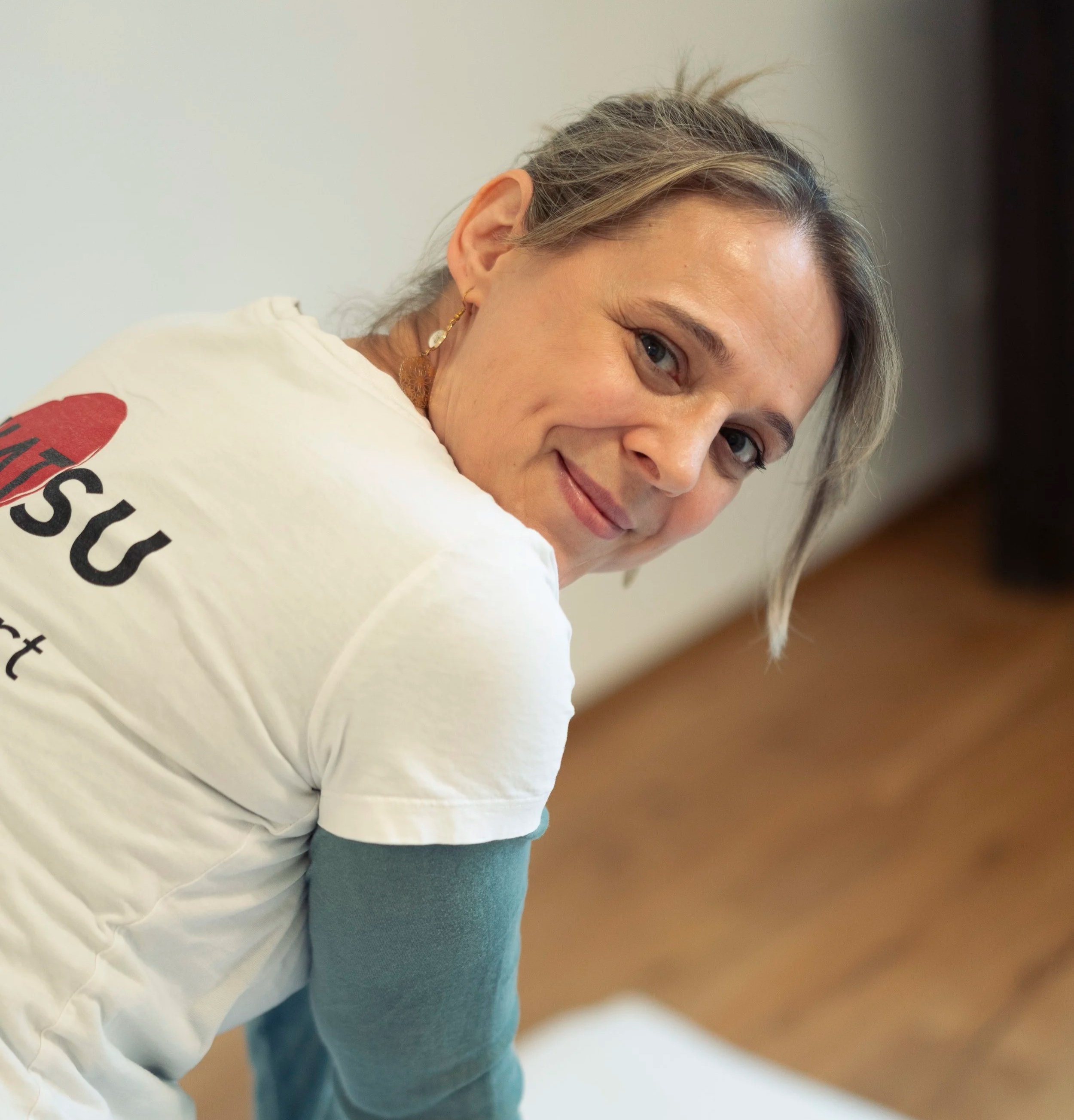 Nahaufnahme eines lächelnden Frauen Gesichts mit blonden Haaren, jeweils ein Ohr mit goldener Ohrring, trägt ein weißes T-Shirt und sitzt auf einem blauen Stuhl.