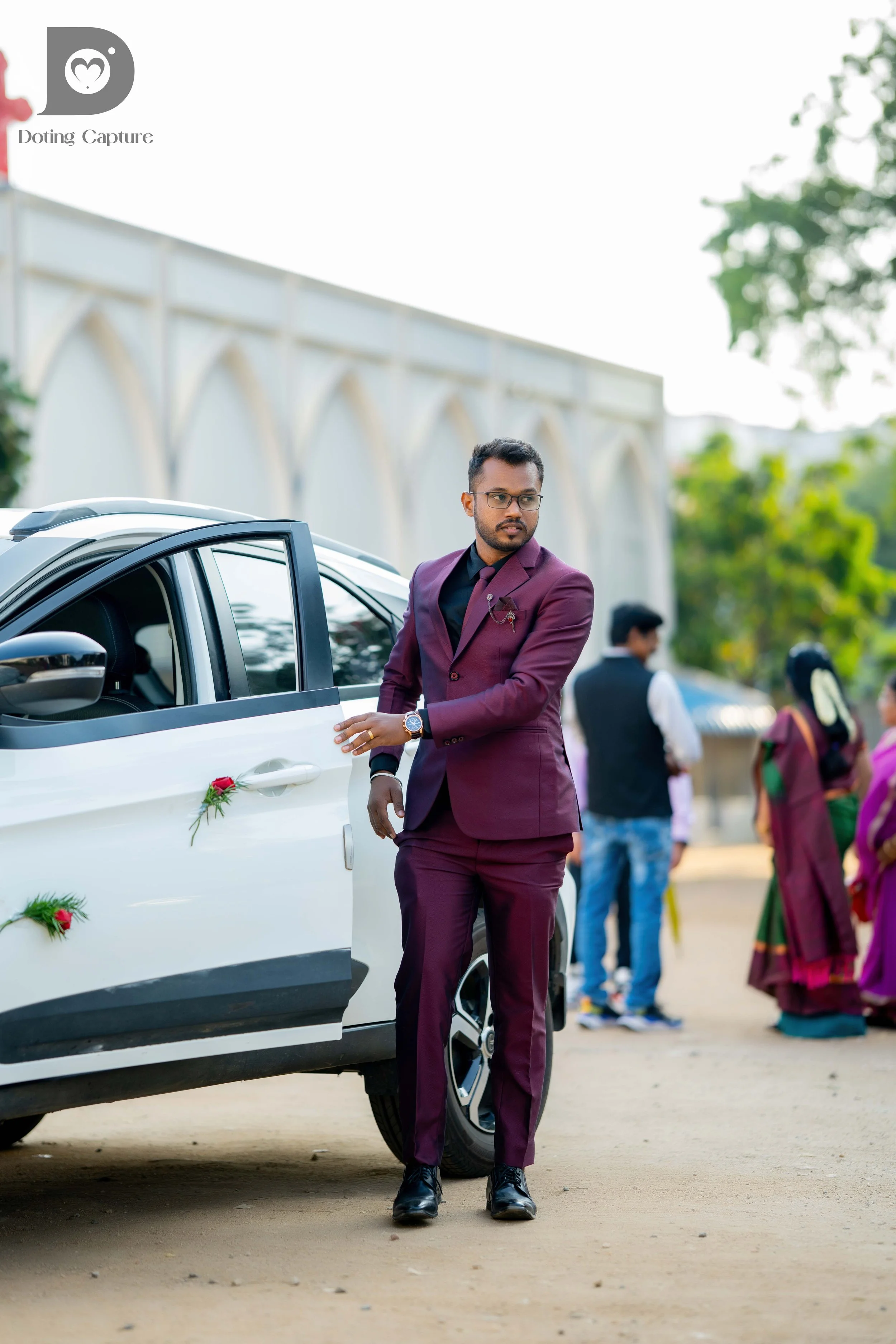 Man in a burgundy suit with glasses and a wristwatch standing next to a white car decorated with small floral arrangements, at an outdoor event with several people in colorful traditional Indian attire in the background.