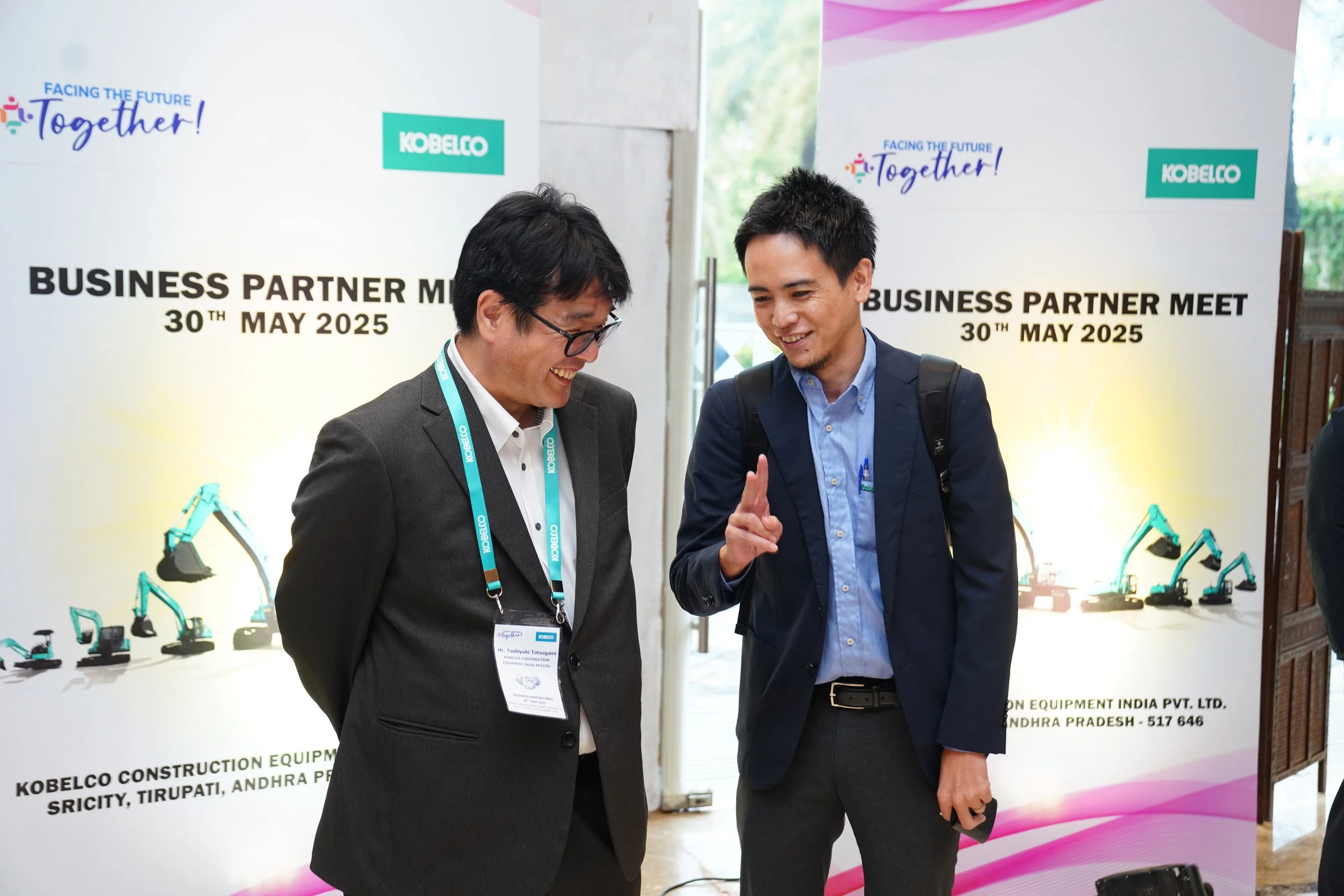 Two men in business attire talking and smiling at a business partner meet event on May 30, 2025, with banners behind them displaying logos and dates.