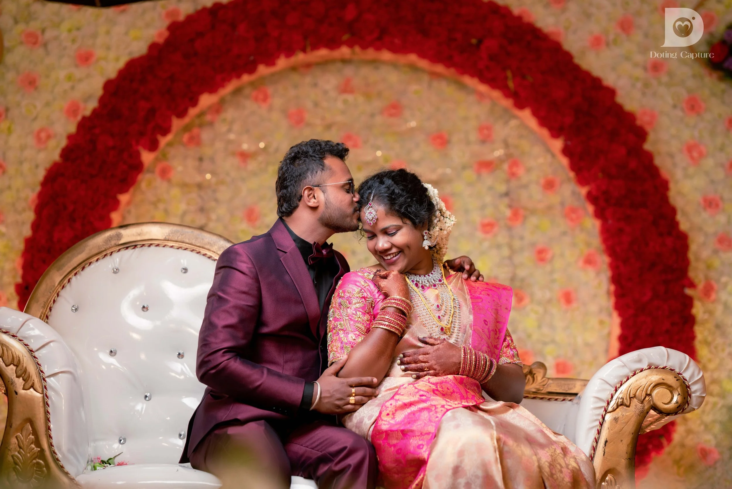 A young couple dressed in traditional and formal attire sitting on a decorated sofa, sharing a tender moment in front of a floral backdrop at a wedding or special event.