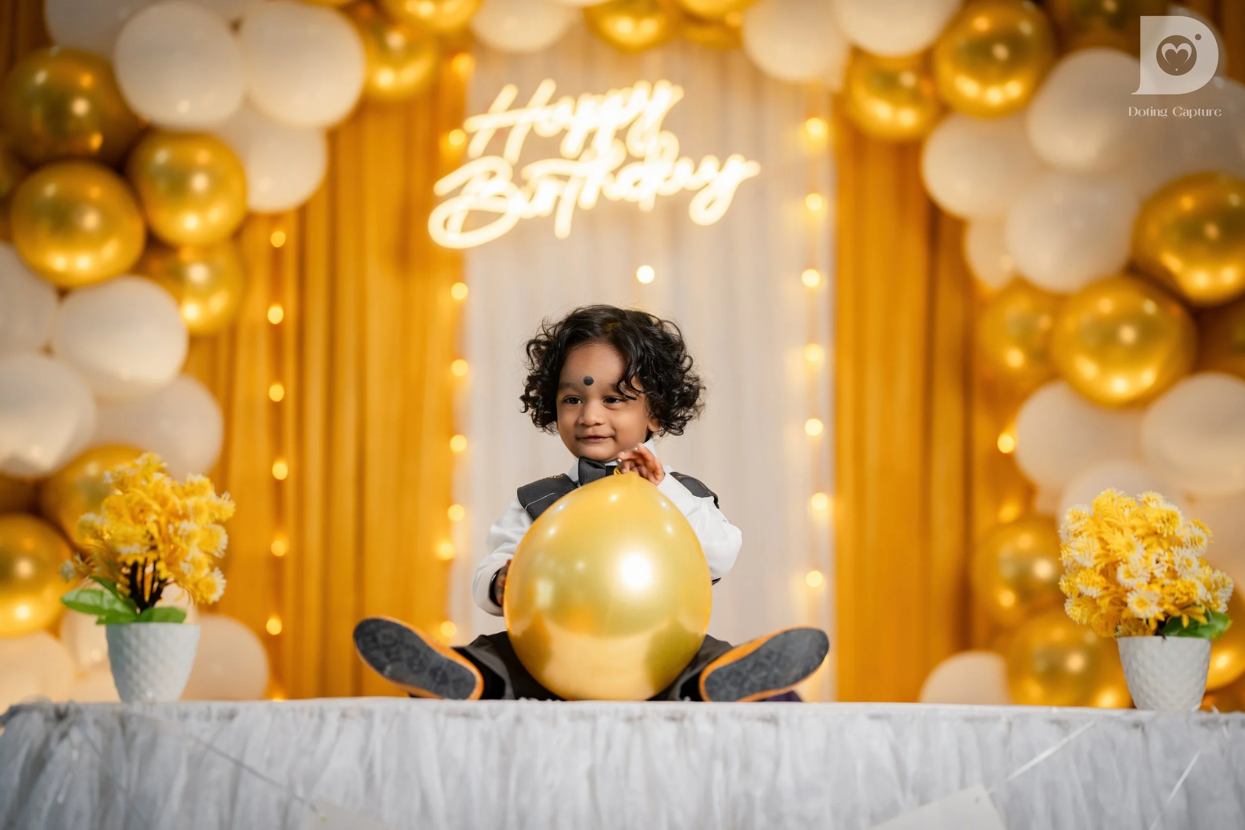 A young child in formal attire sitting on a table holding a large gold balloon, with a decorated background including balloons and a glowing sign that says 'Happy Birthday', flanked by yellow flowers in white pots.