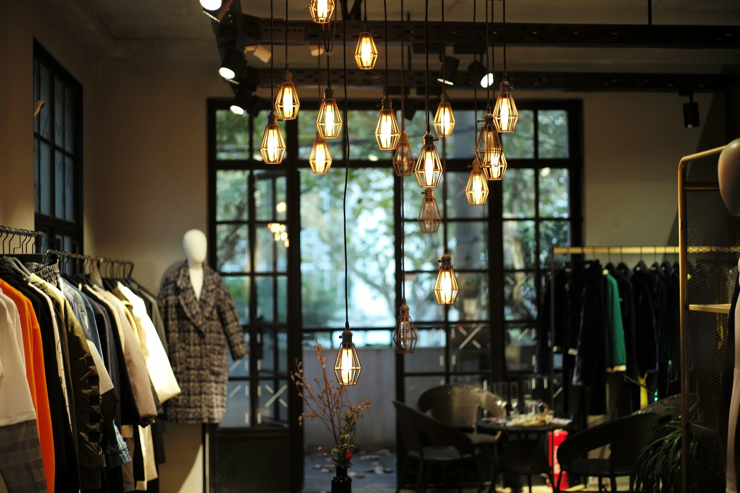 Interior of a clothing store with hanging lights, racks of clothes, and large windows showing outdoor trees.