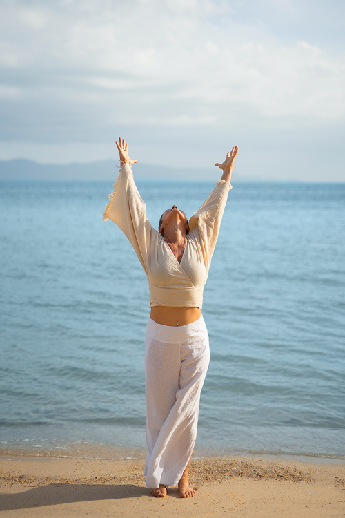 Woman standing on a sandy beach with her arms raised and head tilted back, facing the sky, near the ocean with distant mountains on the horizon.
