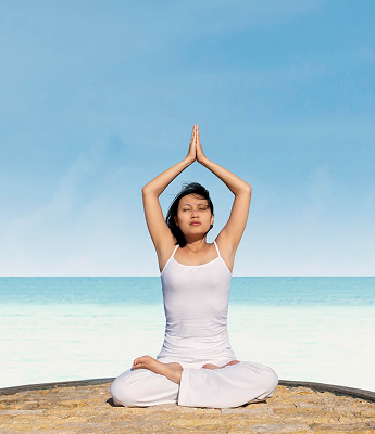 Woman practicing yoga on a beach, sitting in a cross-legged position with hands in prayer pose above her head.