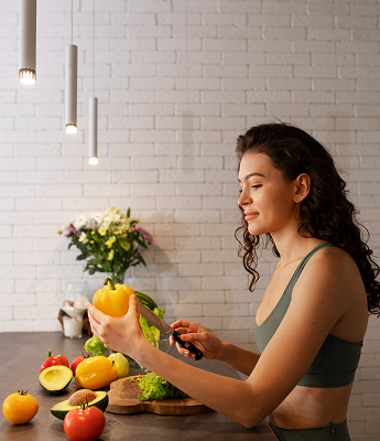 A woman slicing a yellow bell pepper in a modern kitchen with a white brick wall, overhead pendant lights, and a bouquet of flowers in the background.