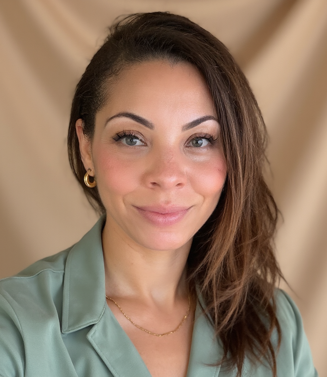 Close-up portrait of a woman with shoulder-length brown hair, blue eyes, wearing gold hoop earrings, a delicate gold chain necklace, and a light green silk blouse, smiling softly against a beige background.