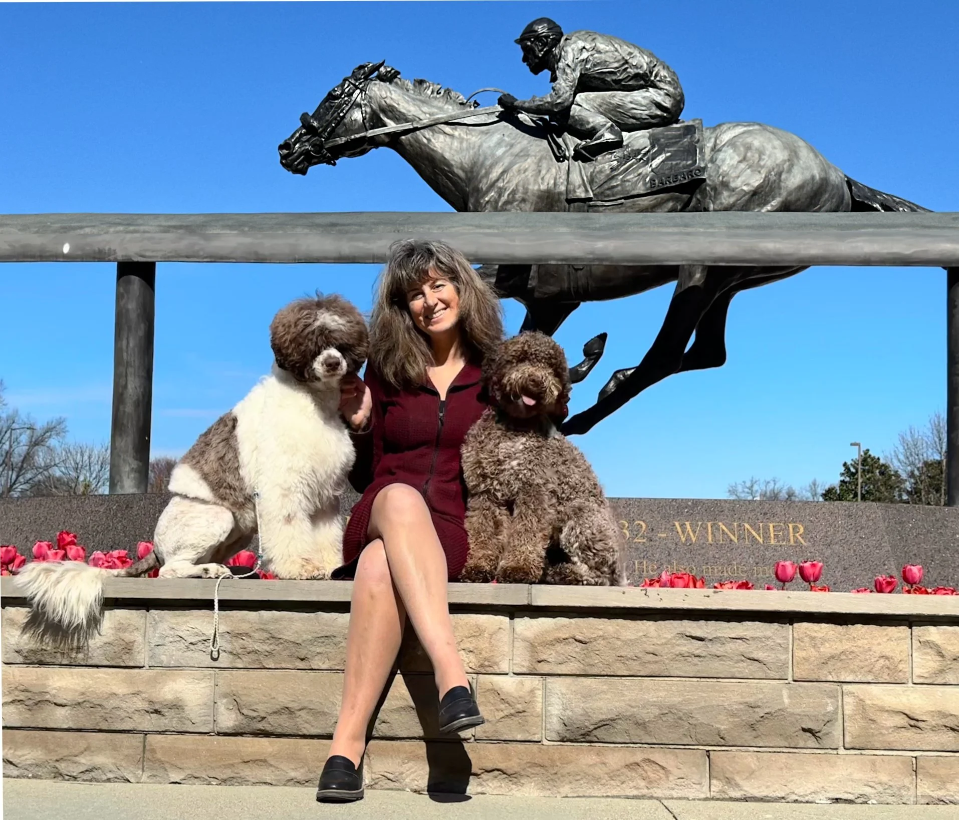 A woman sitting on a stone ledge with two dogs, posing in front of a large equestrian statue and a blue sky. The statue depicts a rider on a horse, and the ledge has red flowers and an inscription reading 'WINNER'.