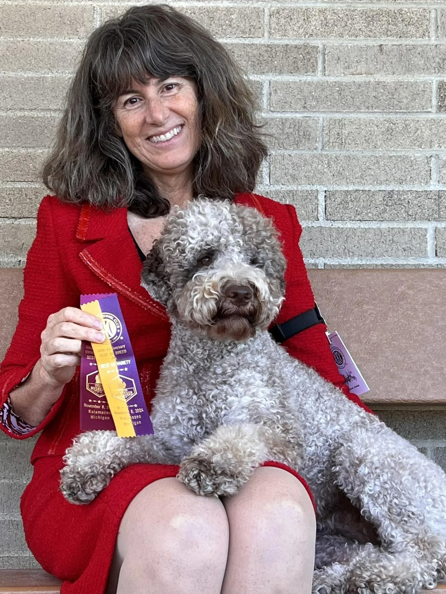 A woman in a red jacket sitting on a bench holding a curly-haired dog with what appears to be ribbon awards.