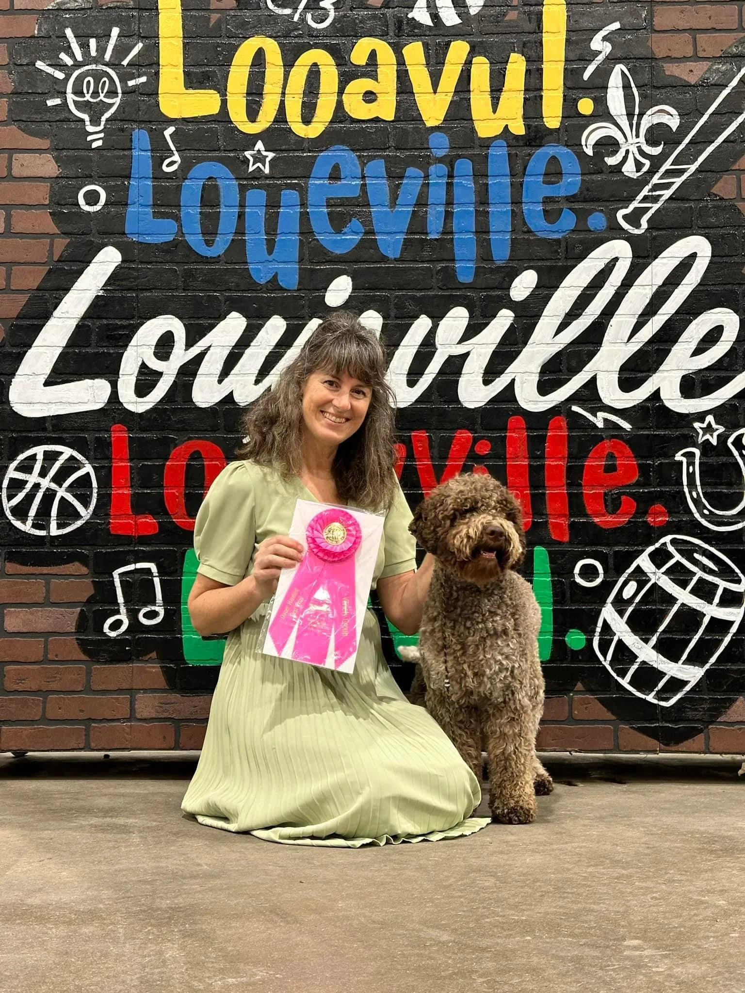 A woman in a light green dress kneels next to a brown curly-haired dog, holding a pink ribbon award. They are in front of a colorful mural that reads 'Loading Louville Louville Louville' with various doodles of a lightbulb, a basketball, musical note