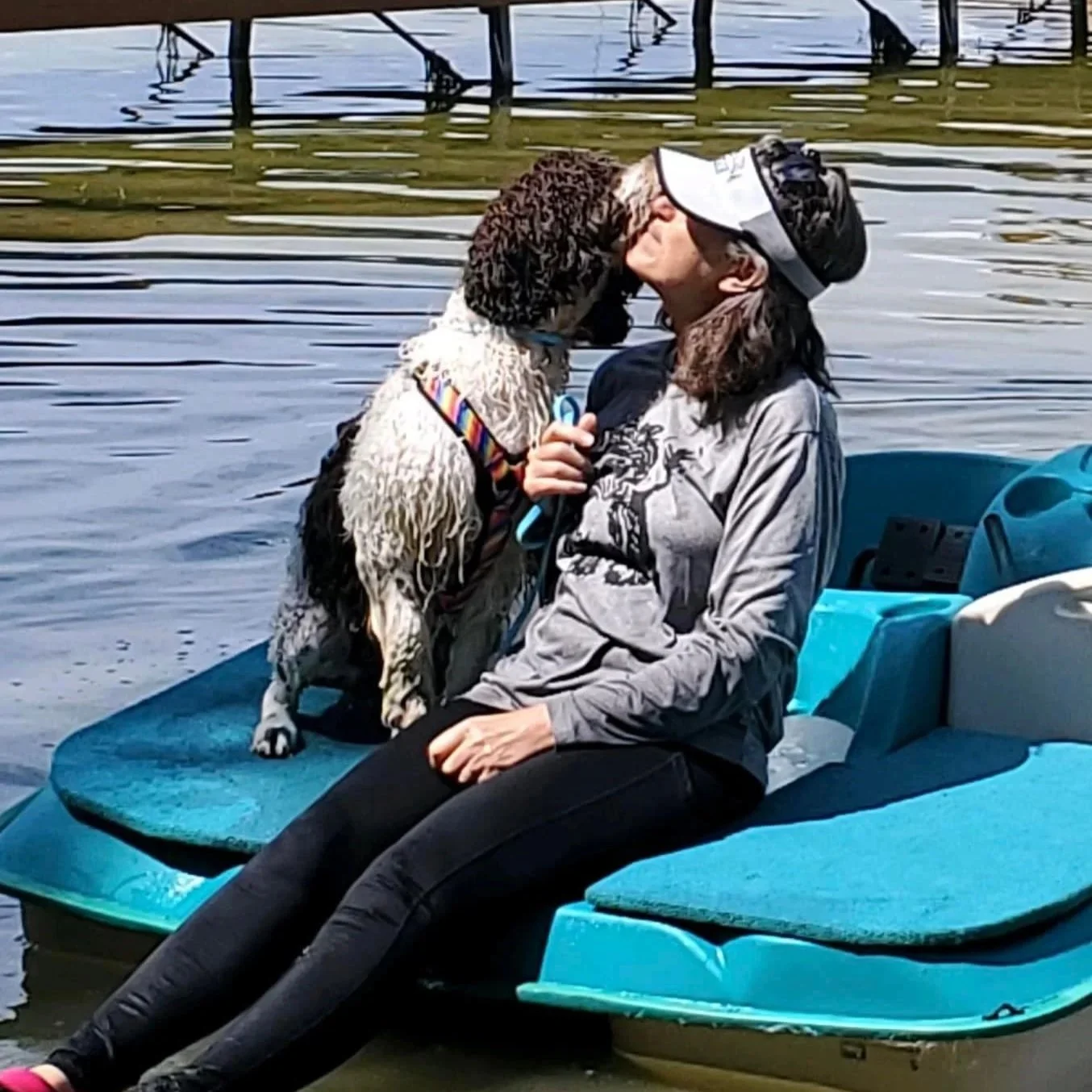 A person sitting on a paddle boat with a dog, kissing the dog on the nose near a dock on a body of water.
